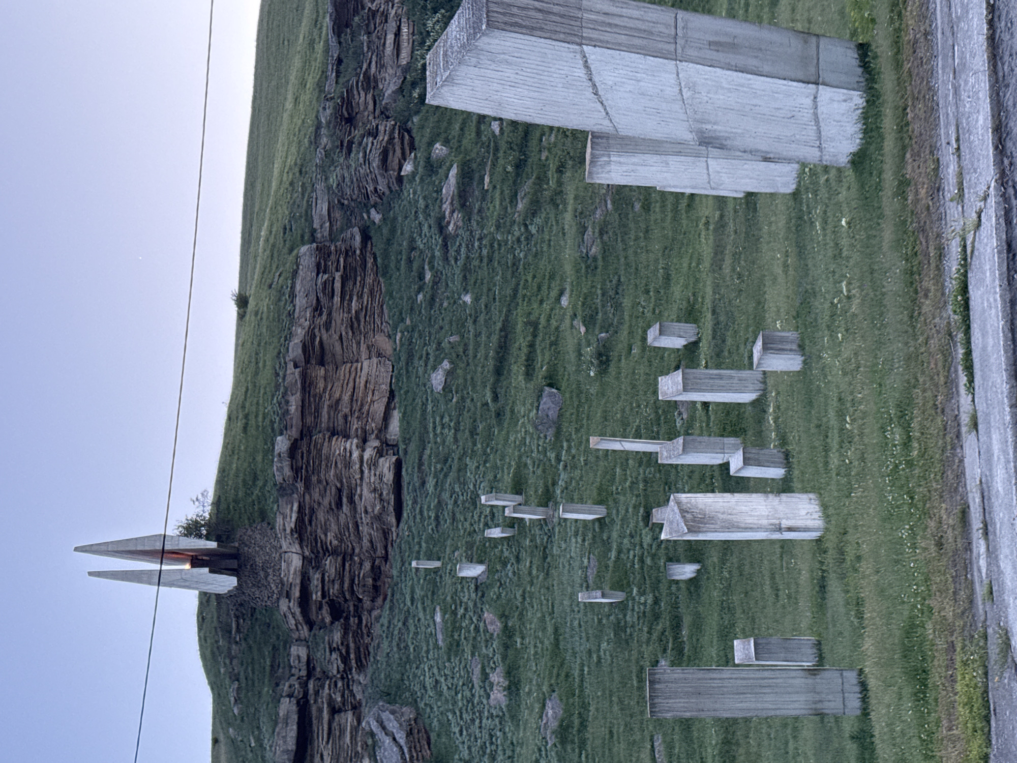The image shows the Memorial to the Victims of Political Repression in Ordzhonikidzevsky, Russia. The memorial consists of a series of rectangular concrete blocks of varying sizes scattered across a grassy hillside, some appearing to jut out from the ground. In the background, there is a rocky cliff face partially covered in vegetation. To the left, a pointed structure with a reddish interior is visible. A road is visible on the right-hand side of the image. A power line runs across the sky. The sky is a light blue color. The grass appears to be green and well-maintained.