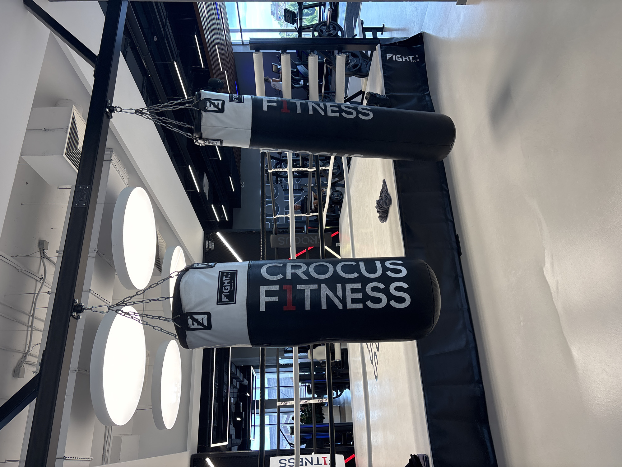 A gym boxing ring features two large, black and white punching bags hanging from a metal support structure. The punching bags have white text that reads "CROCUS FITNESS". The gym has a modern industrial design with exposed ceilings, large circular lights, and sleek black accents. A boxing ring with white padding and ropes is visible in the foreground. In the background, exercise equipment, including treadmills, can be seen. The gym appears to be well-lit and spacious. The image is taken from a low angle, looking up towards the punching bags and the ceiling. The location is identified as Monolit, Russia.