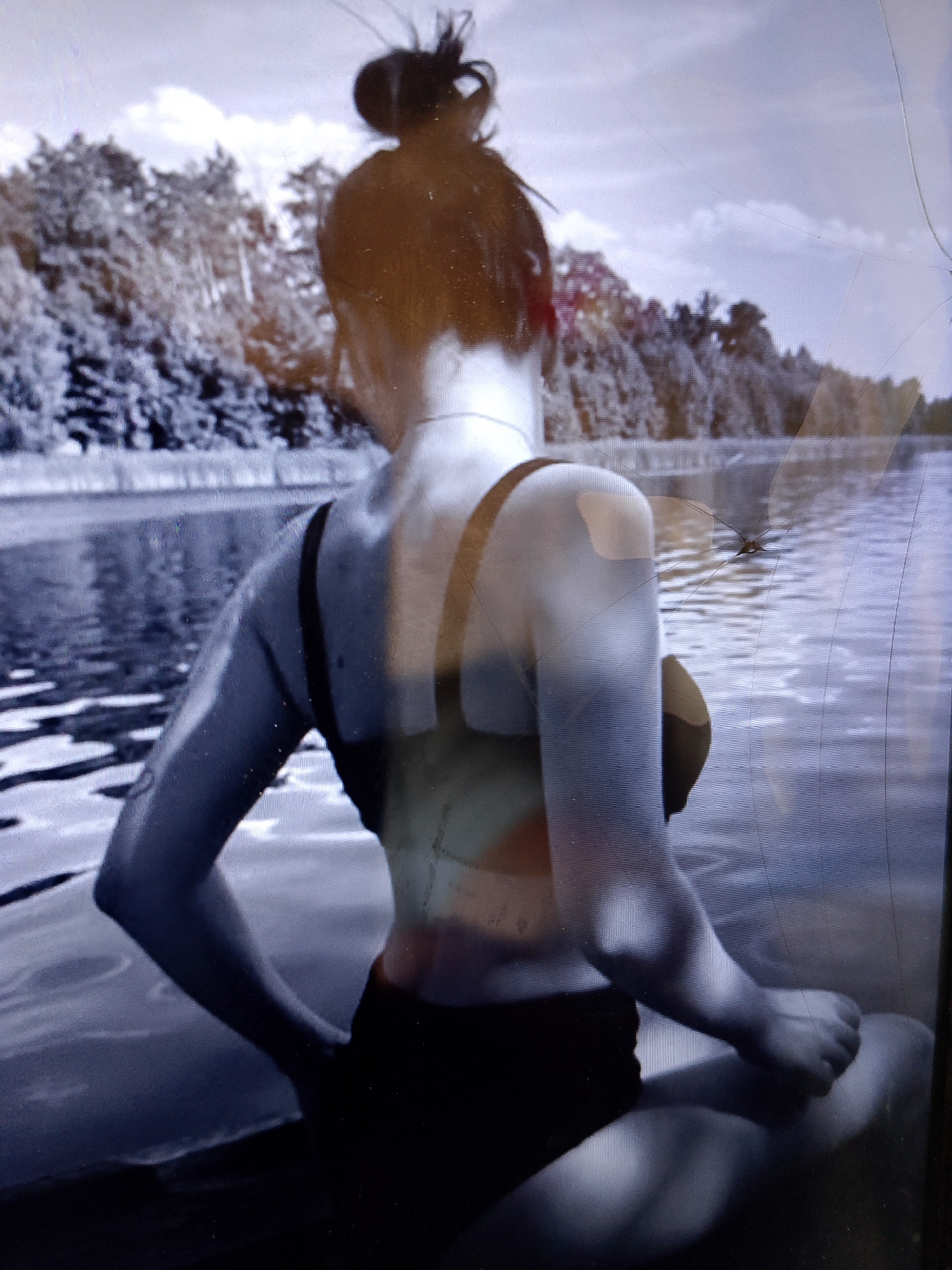 A woman with her hair in a bun is sitting on a body of water, with trees in the background. The image is in black and white, and appears to be taken on a bright, cloudy day. The water is still, reflecting the sky and trees. The woman is wearing a black bathing suit and has her arms resting on her knees. She is facing away from the camera, looking out at the water. The location is in the city of Dobra Krynytsia, Ukraine.