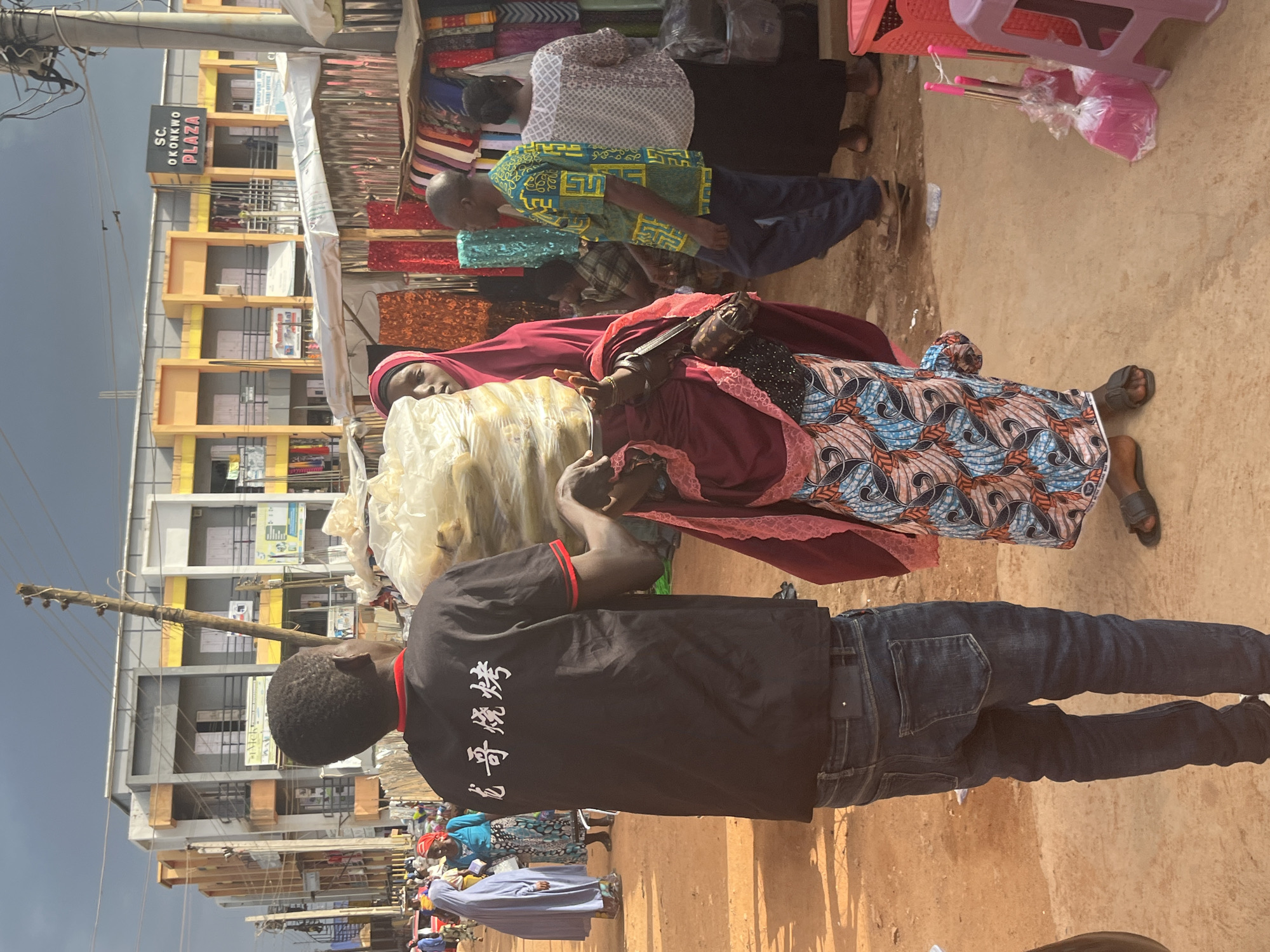 The image captures a bustling scene in Jos, Nigeria, likely a marketplace. In the foreground, a young man wearing a black t-shirt with Chinese characters and blue jeans is seen from the back. He is holding a large, clear plastic bag filled with bananas. To his right, a woman in a red hijab and a colorful patterned dress is also holding the bag of bananas. Behind them, other people are visible, including a woman in a light blue hijab and another in a light-colored patterned top and dark skirt. The background features a multi-story building with an orange facade and the signage "S.C. OKONKWO PLAZA." Stalls with various goods, including fabrics, are set up along the street. The ground is covered in reddish-brown dirt, indicating a dusty outdoor environment. The lighting suggests it is daytime, possibly late afternoon, with the sun casting shadows.