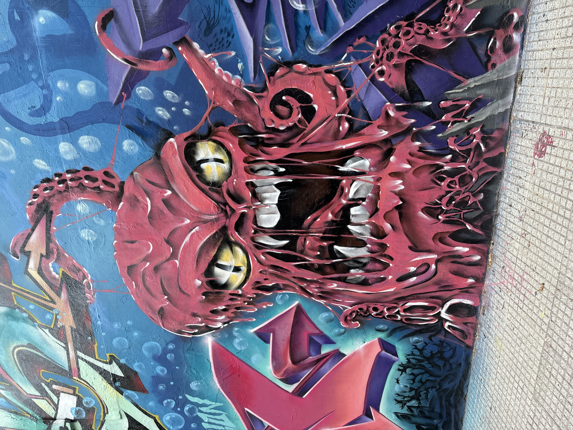 Graffiti covers a wall in Buenos Aires, Argentina. The primary subject is a stylized, monstrous face in shades of pink and red. The face has large, yellow eyes and tooth-like protrusions. Black voids represent the mouth and nose. The background includes blue hues with white bubble-like shapes, purple abstract forms, and sections of geometric shapes. A red and purple arrow-like design is visible at the bottom of the frame. A section of tiled wall is visible on the right.