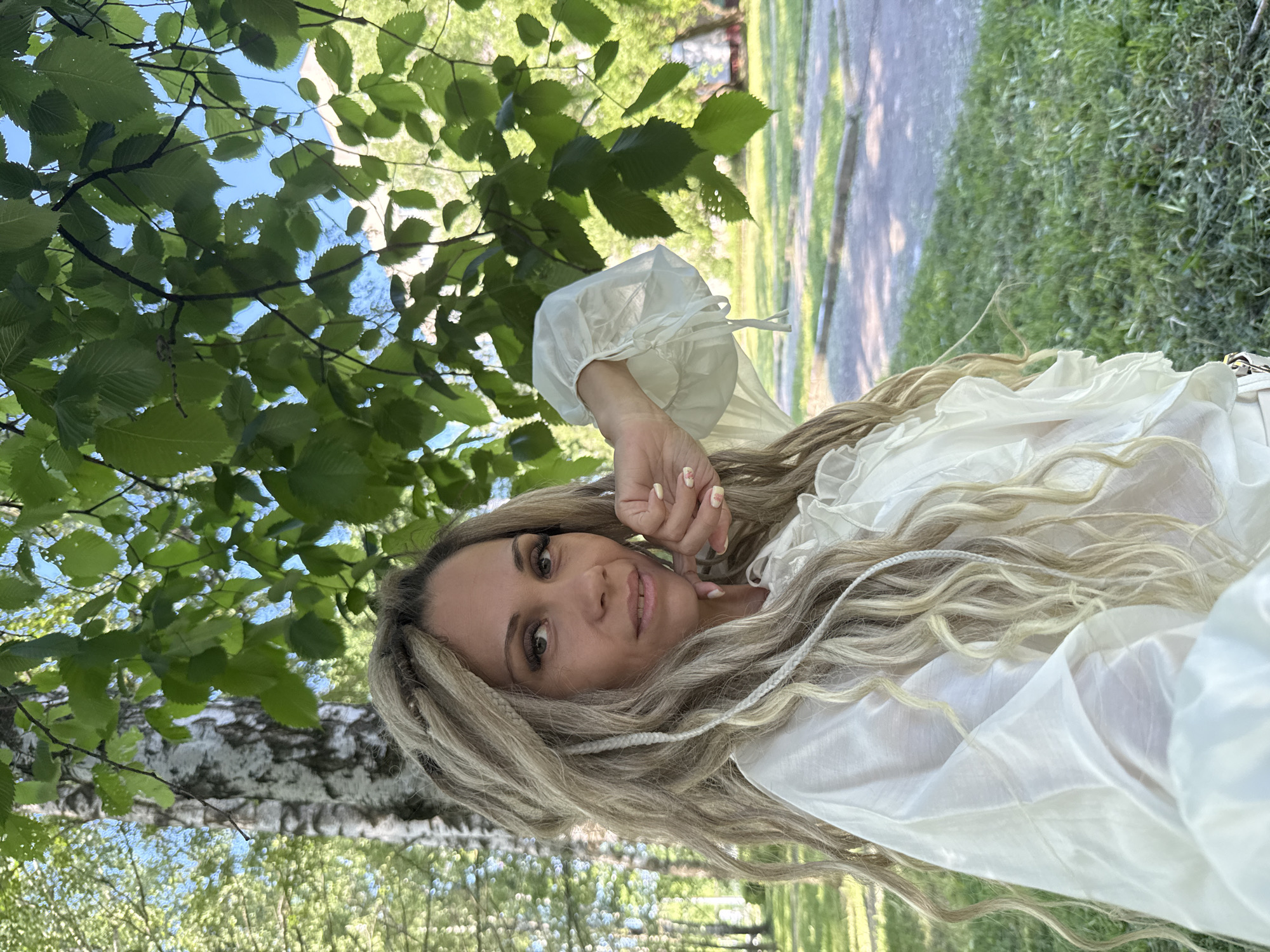A woman with blonde dreadlocks and a white ruffled blouse is pictured outdoors in a park-like setting, possibly Rusanovo, Russia, based on the presence of birch trees and lush green grass. She is looking at the camera with a slight smile and her hand is resting on her cheek. The background shows more trees and a paved area.