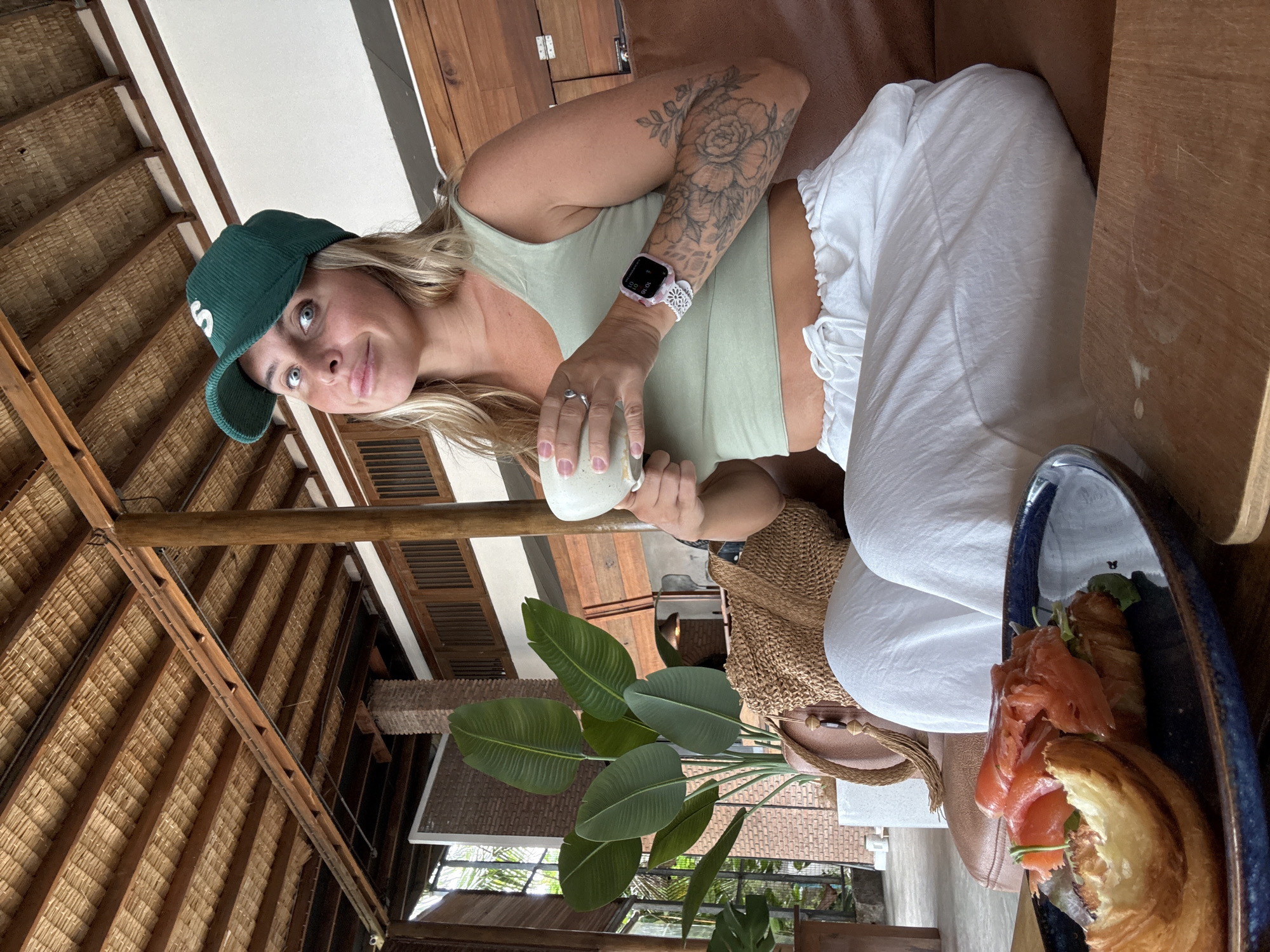 A woman with blonde hair wears a green corduroy baseball cap and a light green crop top. She is seated on a brown leather couch in what appears to be a cafe or restaurant. She holds a white cup in her hands and looks towards the camera with a slight smile. In front of her on a blue plate is an open-faced sandwich with salmon and a croissant. The cafe has a thatched roof supported by wooden beams and pillars. Large potted plants with broad green leaves are visible in the background, along with exposed brickwork and wooden architectural elements. The scene suggests a relaxed, tropical setting, likely in Ko Samui District, Thailand.