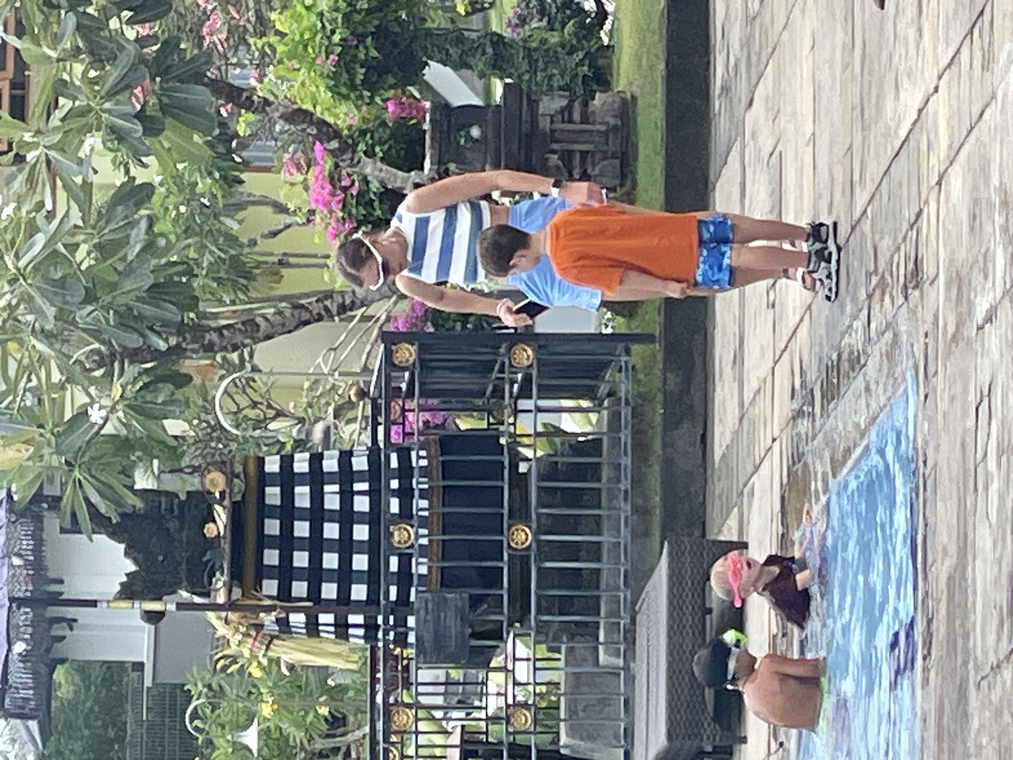In Kuta Selatan, Indonesia, a group of people are near a pool. A woman in a striped crop top and blue shorts stands on a stone patio, looking down at a young boy in an orange t-shirt and blue patterned shorts. In the pool, two young boys are visible. One boy wears goggles and a dark shirt, while another wears a black swim cap and green swim trunks. Behind the people, a decorative structure with a black and white checkered pattern and an umbrella is visible. Lush green trees and pink flowers surround the area, suggesting a tropical resort or garden setting.