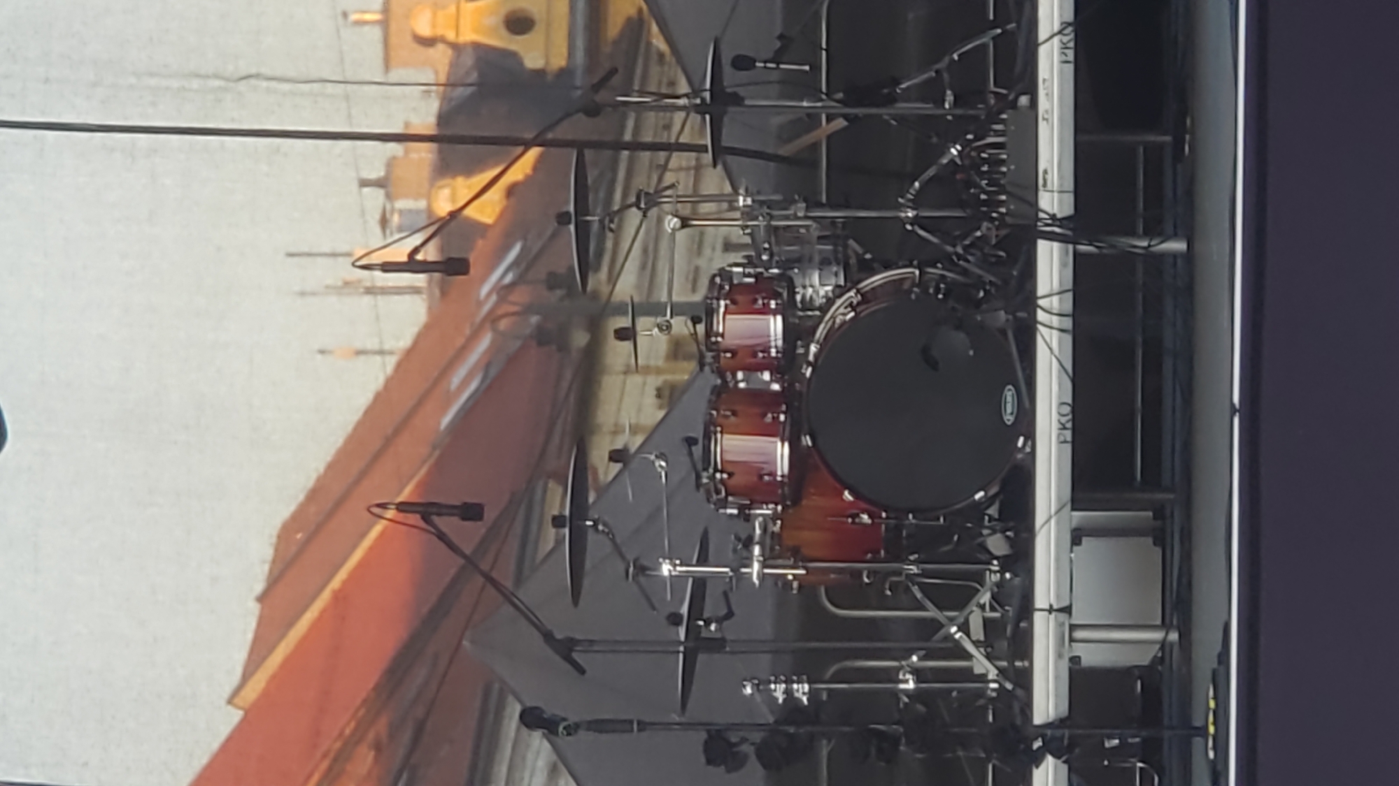 The image shows a drum kit set up on a stage. The drum kit is comprised of several drums and cymbals mounted on stands. Microphones are positioned to capture the sound of the drums. In the background, there is a large projection screen displaying an image of rooftops of buildings in a city, possibly Prešov, Slovakia, with a warm, orange hue, suggesting a sunset or artificial lighting. The stage appears to be outdoors, as indicated by the natural lighting and the open-air environment implied by the background scenery. There are no people visible in the image. The scene is static and appears to be set up for a musical performance. The stage structure has the logo "PKO" visible on its side.