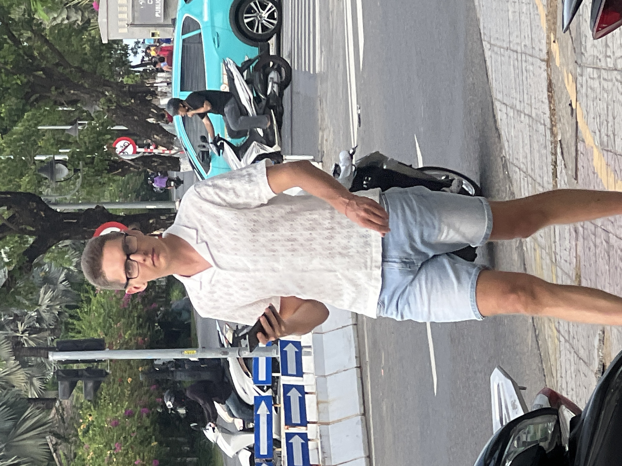 The image captures a street scene in Da Nang, Vietnam, with a young man in the foreground walking while looking at his smartphone. He is wearing a white patterned short-sleeved shirt and light blue denim shorts, along with glasses. Behind him, traffic signals are visible, and several motorbikes are present, some being ridden by other individuals. A bright teal car is also in motion on the road. The background features trees, foliage, and a building, suggesting an urban environment. White and blue directional signs with arrows are mounted on a low concrete barrier on the side of the road. The pavement is paved with interlocking bricks, and there is a yellow line demarcating a pedestrian walkway.