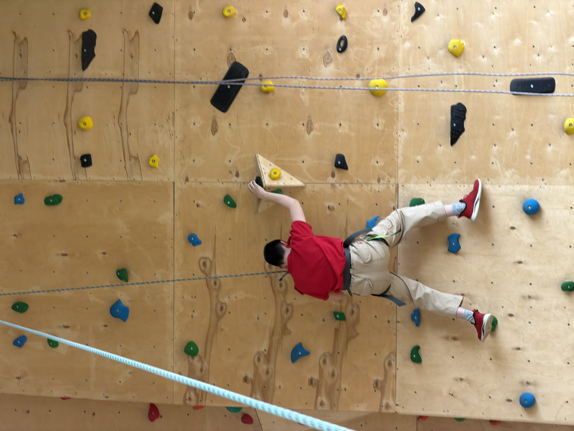 An indoor rock climbing wall in Izhevsk, Russia, is depicted from a slightly elevated perspective, showcasing a person actively engaged in climbing. The wall is constructed from numerous light-colored plywood panels, with visible screw holes and wood grain. A variety of rock climbing holds in different shapes, sizes, and colors—including yellow, black, blue, green, and red—are affixed to the wall, alongside a large, prominent triangular wooden block hold.
A person, likely a youth, is captured mid-climb, oriented horizontally across the wall. They are dressed in a red long-sleeved shirt, beige or khaki pants, and red sneakers with gray socks, and are wearing a climbing harness. Their body is extended and appears to be under tension, demonstrating the physical effort of the activity. The right arm is outstretched, gripping the large wooden triangular hold, while the left arm is bent, reaching for another hold. The left foot is placed on a green climbing hold, and the right foot is on a black hold, with a blue hold nearby.
Two ropes, one blue and white, and the other purple and white, are strung horizontally across the climbing wall at different heights. These ropes appear to be part of the climbing system, possibly marking routes or for an auto-belay. The scene takes place in an indoor facility, indicated by the artificial lighting and plywood construction. There are no specific external weather or time-of-day cues. No visible text is present in the image. The picture captures a dynamic moment of focused physical activity within a dedicated climbing environment.