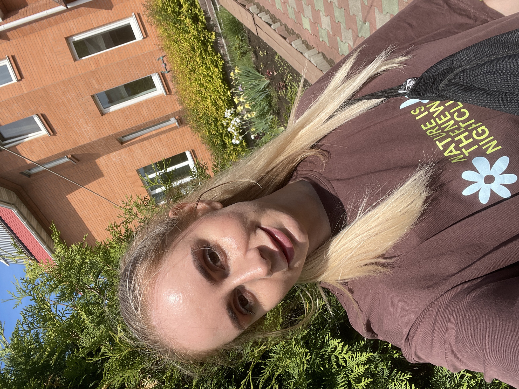 A blonde woman takes a selfie outdoors. She wears a brown sweatshirt with a graphic flower and text that reads "NATURE IS THE NEW NIGHTCL". A black backpack is visible over her shoulder. A brick building with white-framed windows is in the background, along with green landscaping. The ground is covered in red and green pavers. Green foliage is in the foreground. Based on the location metadata, the image was taken in Vladivostok, Russia.