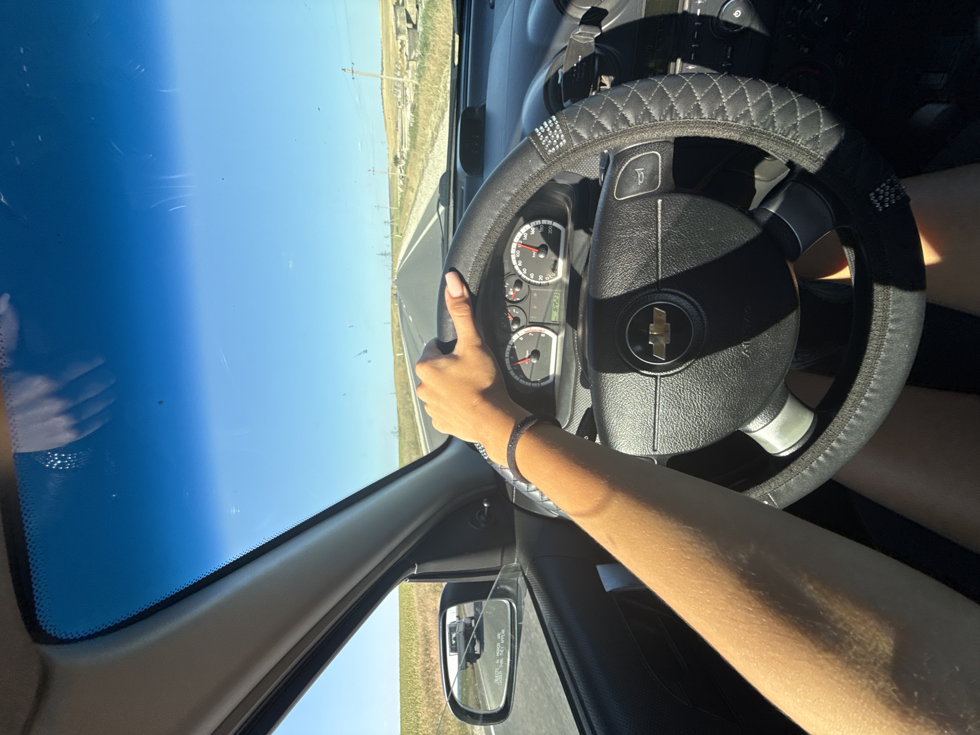 The image shows a first-person perspective from inside a car traveling on a straight, paved road. The driver's arm, tanned and wearing a dark bracelet, is visible on the steering wheel, which is covered with a black, quilted material and adorned with rhinestones. The dashboard displays the car's speed at approximately 110 km/h and an odometer reading of 164523. The road is bordered by dry, grassy fields, with scattered utility poles and some remnants of concrete structures. The sky is clear and blue. In the side mirror, another vehicle, possibly a truck, is visible on the same road. The sun appears to be shining brightly, casting shadows within the car. The scene is set during the daytime.