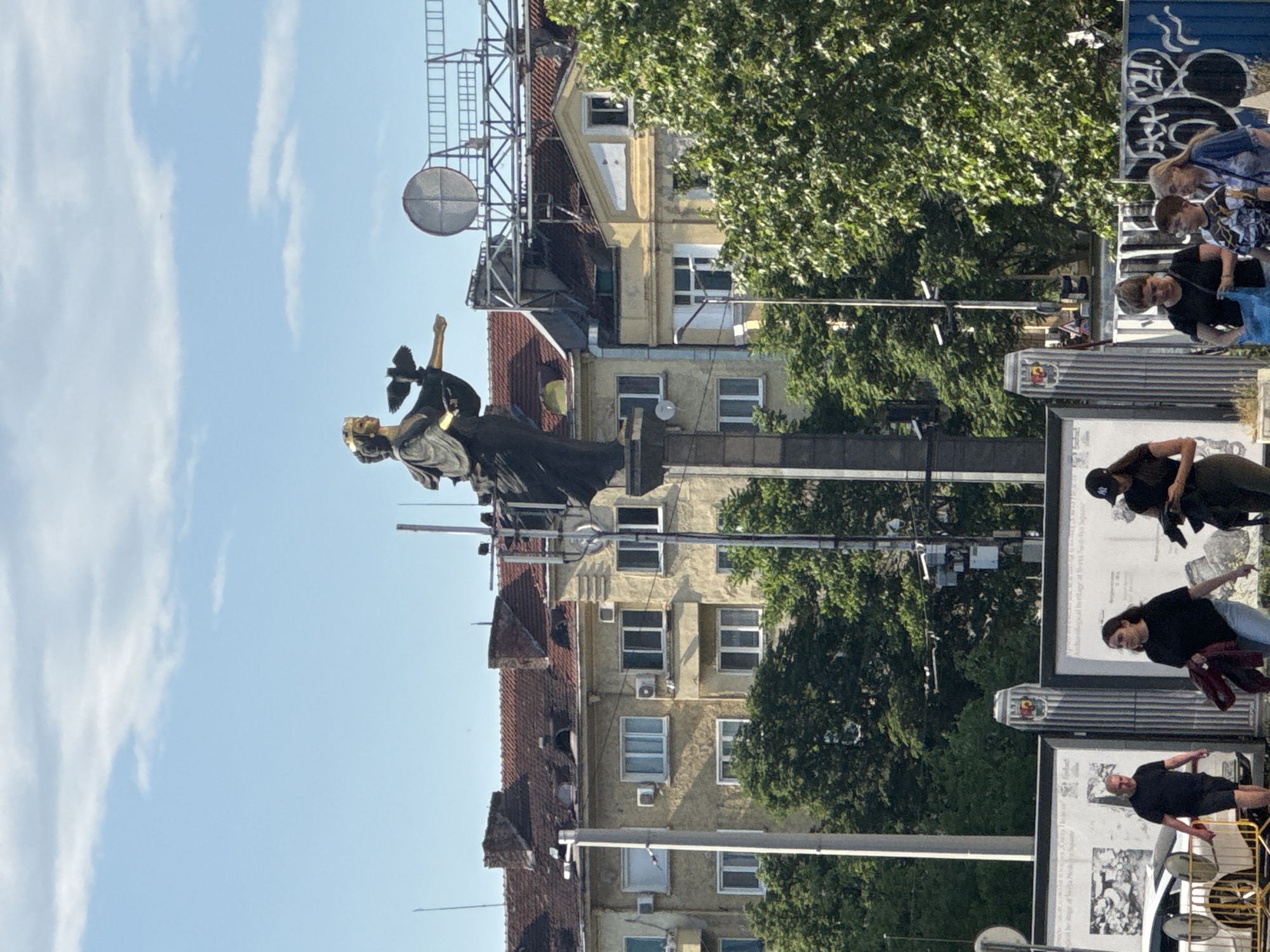 The image depicts a vibrant urban street scene in Sofia, Bulgaria, bathed in bright sunlight under a clear blue sky with scattered clouds.
In the left-mid ground, a prominent, tall statue of a female figure is visible. She wears a crown or ornate headdress and appears to be holding an object, possibly a bird or a branch, in her raised hand. The statue, dark in color (likely bronze or dark stone) with golden accents, stands atop a tall, dark pedestal.
Behind the statue and extending across the mid-ground are several multi-story buildings, characterized by their terracotta tiled roofs and light-colored (yellow/beige) facades. These buildings feature numerous windows, some equipped with air conditioning units. Scaffolding is visible on the upper part of one building, suggesting ongoing maintenance or construction. Lush green trees are interspersed throughout, indicating a warm season.
On the right side of the image, several individuals dressed in casual attire are present, some seemingly observing large, vertical informational display boards. These boards contain black and white images and partially legible text, including phrases such as "located in the very center of Sofia" and references to "Nezavisimost Square," confirming the geographical context. Graffiti marks a blue utility box or wall section. Various urban elements like lampposts, overhead wires, and a large parabolic dish mounted on a structure further populate the scene, contributing to the bustling city atmosphere.