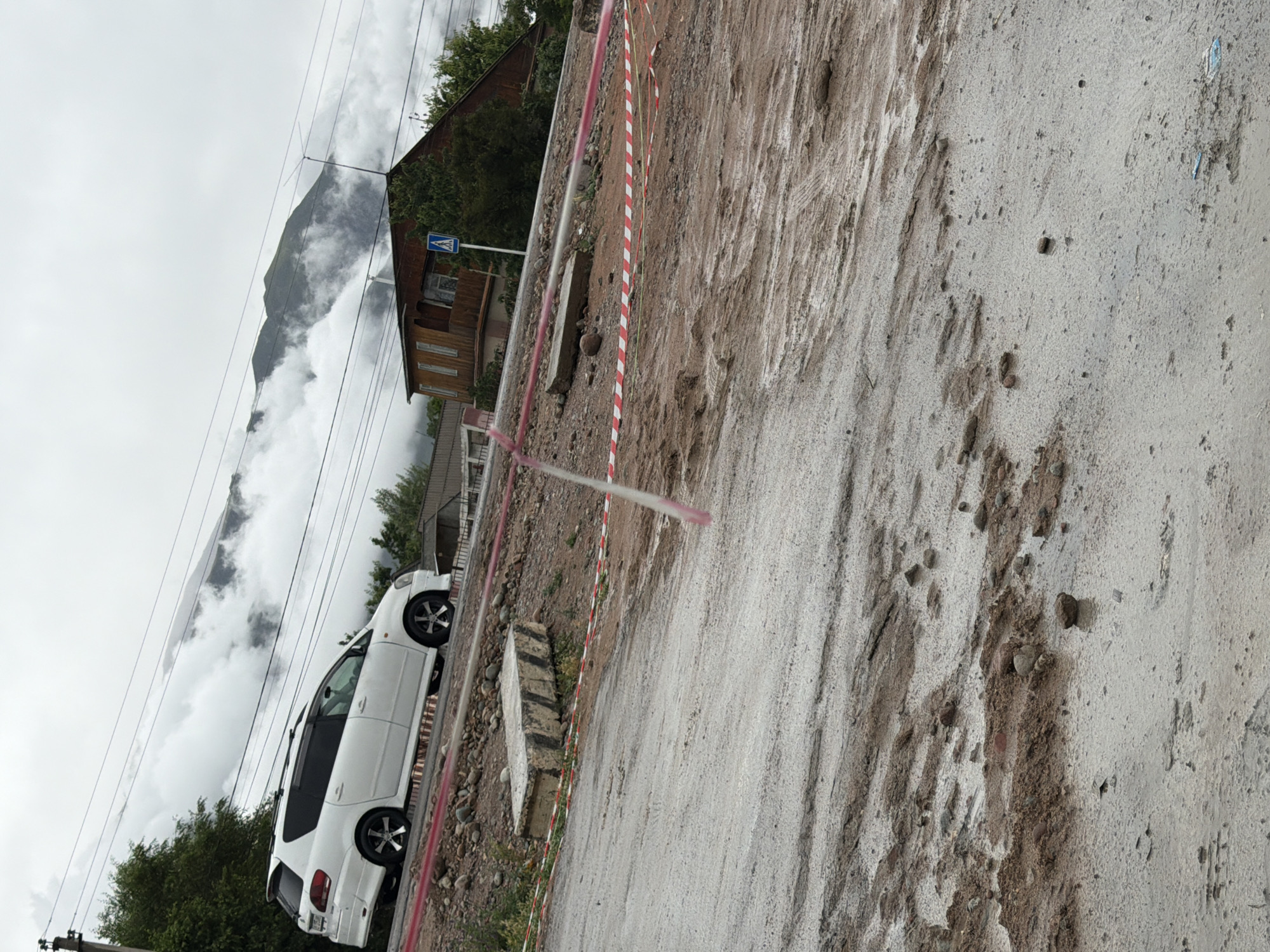 The image depicts a muddy, partially constructed or damaged road in the city of Kara-Oy, Kyrgyzstan. In the background, a white minivan is parked on the side of the road. Beyond the minivan, a brown wooden house with a balcony is visible, alongside a pedestrian crossing sign. The road surface is covered in mud, gravel, and debris, indicating ongoing construction or a recent weather event. Red and white caution tape is strung across the road in several sections, warning of the hazardous conditions. The sky is overcast, and mountains shrouded in clouds are visible in the distance. The scene suggests a rural or semi-rural setting within Kara-Oy.