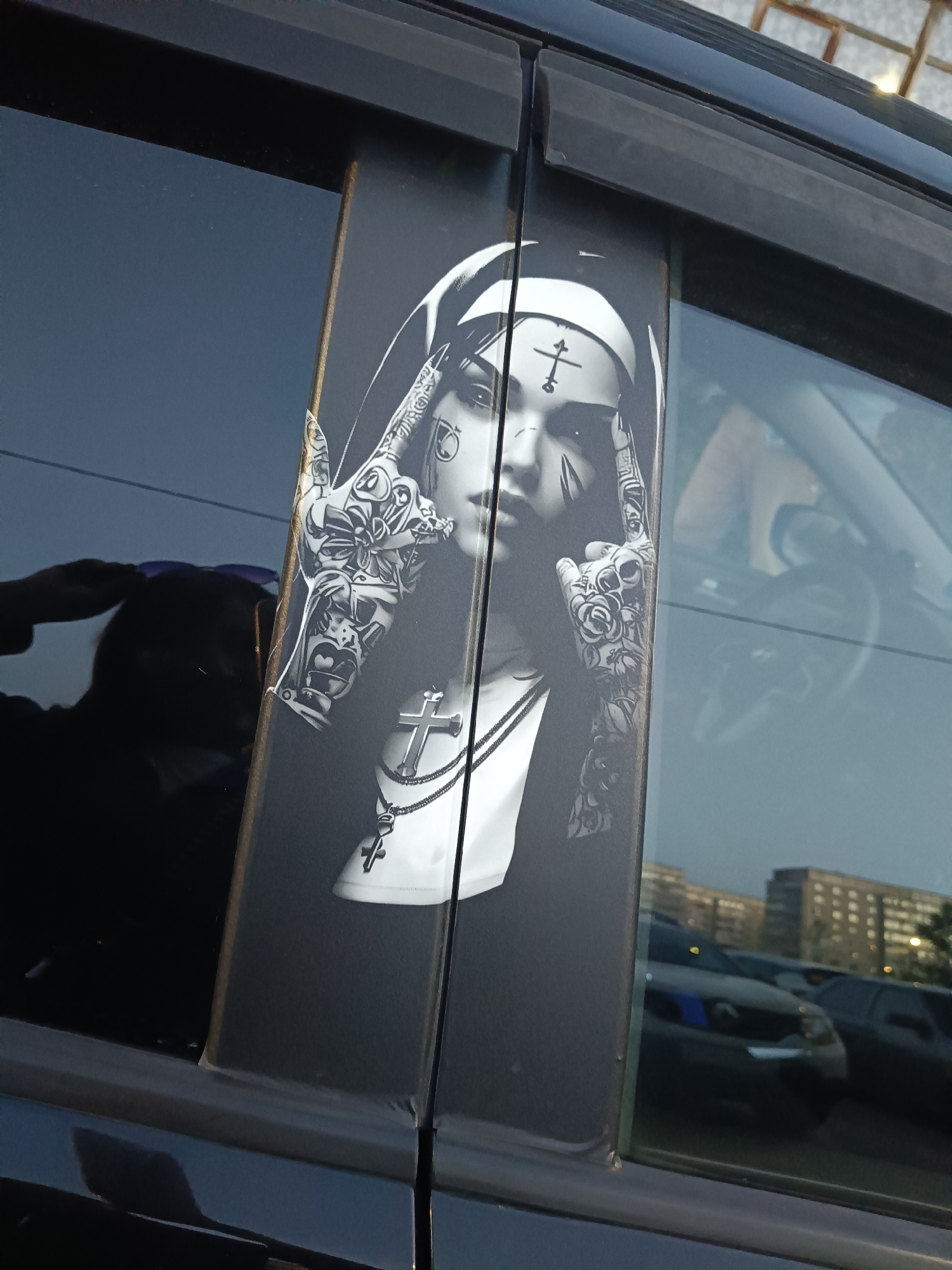 The image shows a close-up view of a car door with a sticker of a stylized nun. The sticker is in black and white and depicts the nun with tattoos on her hands and face, and a cross necklace. The nun's head is tilted slightly, and her hands are raised to her temples. A reflection of a person can be seen in the car window, and the background shows buildings and other cars, suggesting an urban environment. The photographer's camera is held at eye level, looking slightly downwards at the sticker. The lighting suggests it is daytime, possibly late afternoon or early evening. No specific location in Yoshkar-Ola, Russia, can be identified from the image itself.