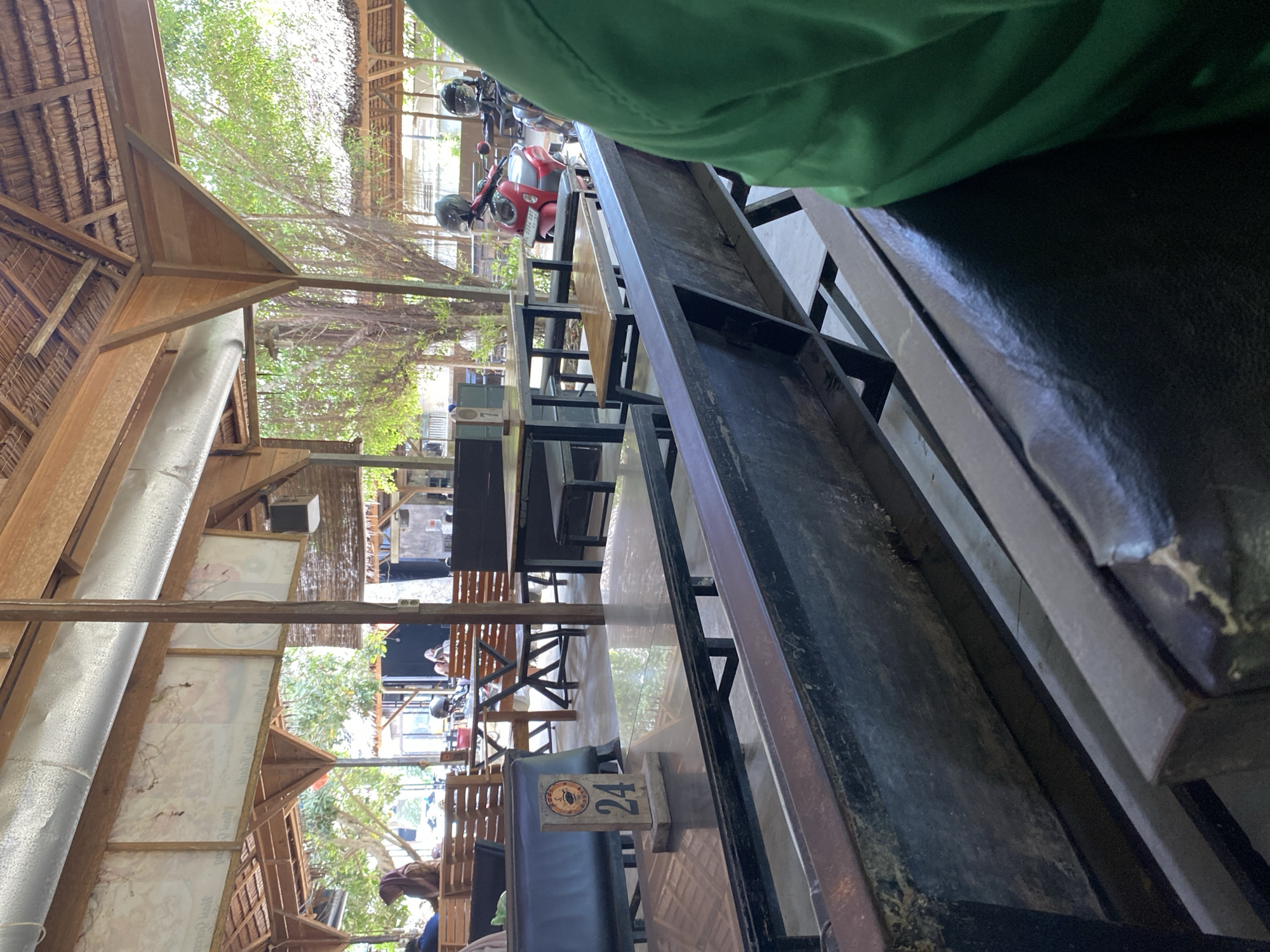 The image depicts an outdoor seating area, potentially a cafe or restaurant, in Gampongaceh, Indonesia. The structure features a thatched roof supported by wooden beams and pillars. Tables and benches are visible, constructed of metal and wood. Table number "24" is displayed. Several motorcycles are parked in the distance. The environment suggests a tropical or semi-tropical climate, with visible greenery.