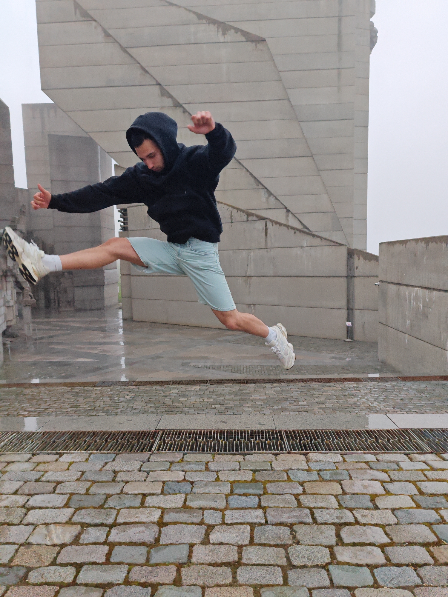 A man is captured mid-air in a wide stance leap. He wears a dark hoodie, light blue shorts, and white athletic shoes. His left leg is extended forward, while his right leg extends behind him. His right arm is raised, with a fist clenched, and his left arm is extended to the side with his thumb pointing up. The background features the Founders of the Bulgarian State Monument in Shumen, Bulgaria, a large, gray, brutalist architectural structure. The ground appears to be wet, with various stone surfaces and a metal drainage grate in the foreground. The day appears overcast or rainy.