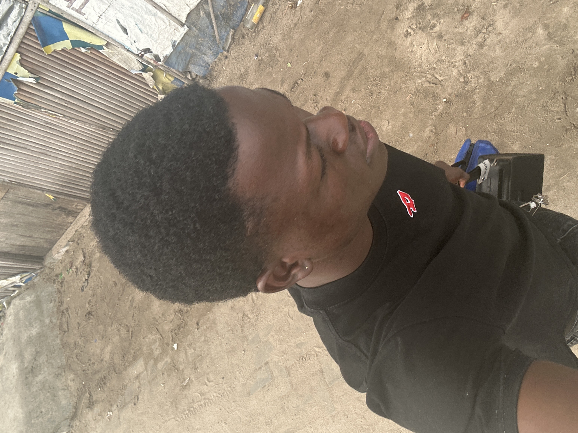 An overhead shot captures a young Black male from the chest up, angled to his right, standing on a sandy, dirt path. He wears a black t-shirt with a small red and white logo on the left chest and dark pants. He has short, dark, curly hair and is wearing a small silver stud earring in his left ear. In his right hand, he holds a black rectangular object with a blue strap attached, possibly a bag or case, which is partially obscured by his shirt.
The background consists of a weathered, corrugated metal fence, likely part of a structure or barrier, with pieces of ripped blue and yellow fabric or posters attached to it. To the left of the fence, the ground is uneven and dusty, with scattered debris. The lighting suggests it is daytime, with sunlight casting shadows. The scene appears to be in an outdoor, somewhat undeveloped area, consistent with observations of the outskirts of Badagry, Nigeria, where such fencing and ground conditions are common.