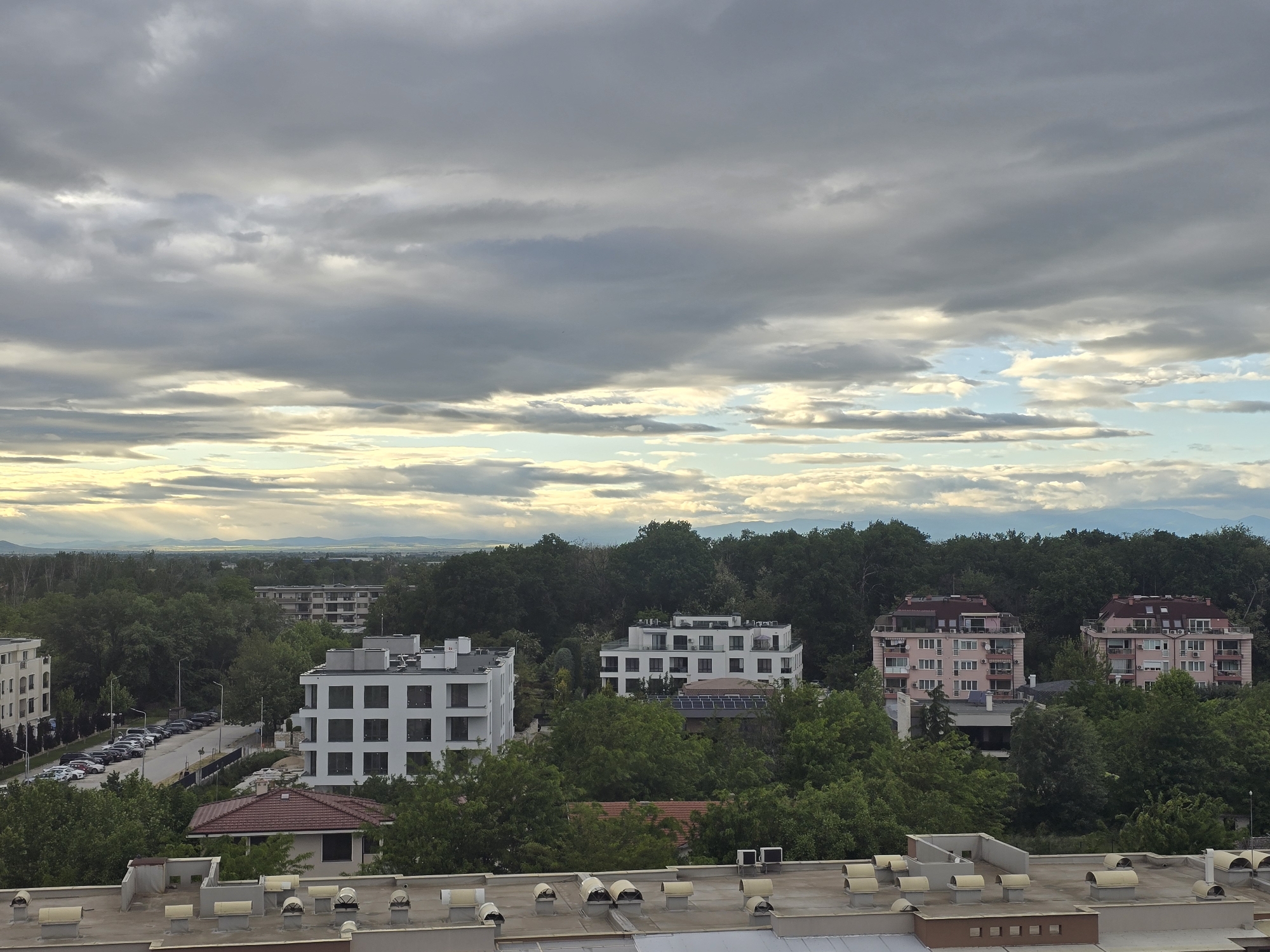 The image depicts an urban landscape in Unknown, Unknown, featuring a rooftop view overlooking a neighborhood. The sky is overcast with a mix of dark gray and lighter, sunlit clouds. Below, a series of residential buildings of varying heights and architectural styles are visible, including modern white structures and older pinkish buildings. The buildings are interspersed with lush green trees, creating a dense urban forest. A street with parked cars is seen on the left side of the image. In the distance, a faint skyline suggests hills or mountains. The foreground shows a flat rooftop with numerous air conditioning units.