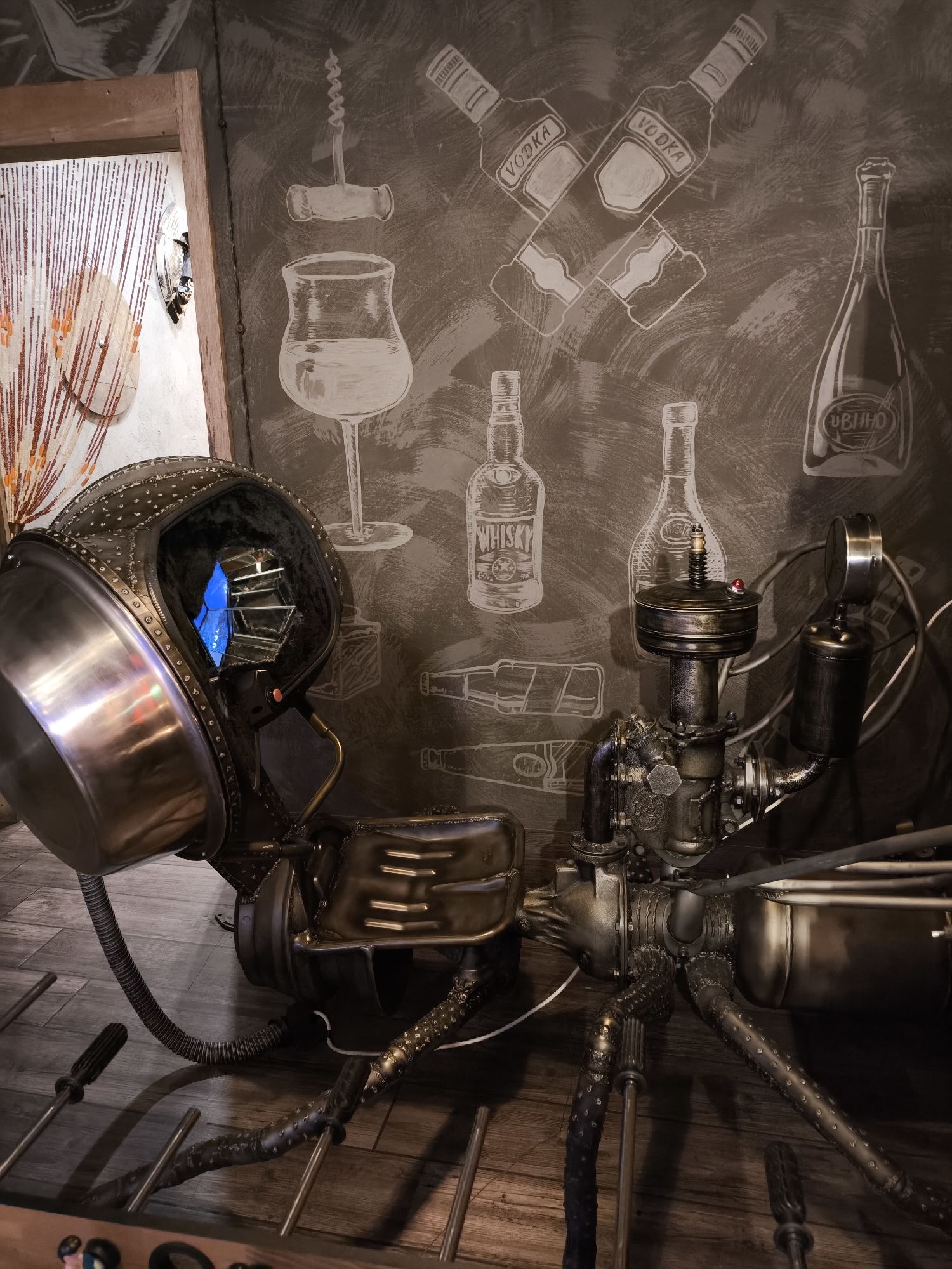This image depicts a close-up, eye-level view of a steampunk-inspired robotic sculpture, likely located in Yekaterinburg, Russia. The sculpture features a large, metallic, helmet-like head with a blue light emanating from within, resting on a complex mechanical body with multiple articulated limbs resembling insect legs. The sculpture is positioned in front of a wall adorned with white chalk drawings of bottles, a wine glass, and a corkscrew, suggestive of a bar or restaurant setting. The wall has a textured, dark brown finish. The floor is made of wooden planks. To the left, a doorway with beaded curtains and a glimpse of an interior space is visible. The overall aesthetic is dark, industrial, and artistic, with a focus on intricate mechanical details and retro-futuristic design elements.
