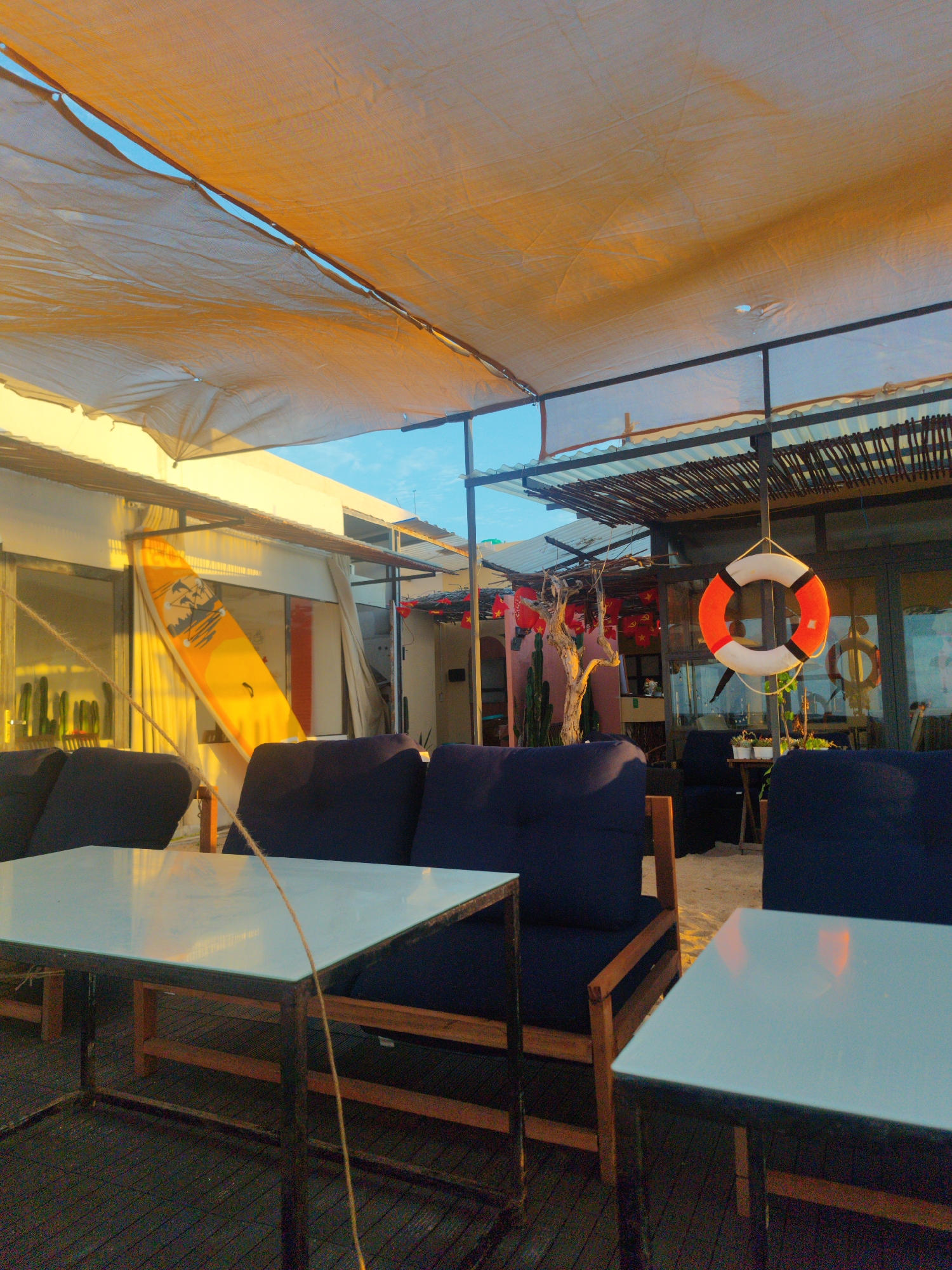 The image depicts an outdoor seating area, possibly a restaurant or lounge, in Phú Quý Special Administrative Region, Vietnam. The scene features several dark blue upholstered seating arrangements with light-colored tabletops. A yellow surfboard is leaning against a building. A life preserver hangs in the background near a tree decorated with red lanterns and cacti. Overhead, a sunshade is stretched across a metal frame, allowing some sunlight to filter through. The ground appears to be wooden slats in the foreground and sand in the background.
