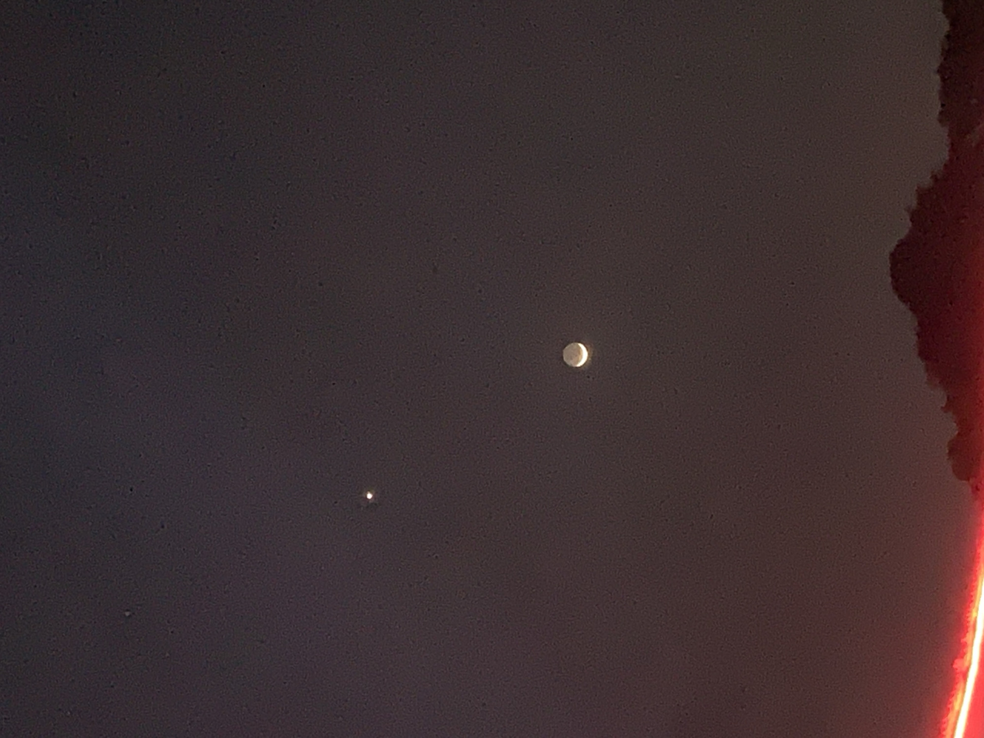 The image shows a night sky over New Stadium, Nigeria. A bright, nearly full moon is visible in the upper center of the frame. A smaller, fainter celestial object, possibly a planet or a bright star, is located below and to the left of the moon. The sky is dark, suggesting a nighttime setting. A blurred, reddish object, likely out of focus, is present along the right edge of the image. No people or other objects are identifiable in the image.