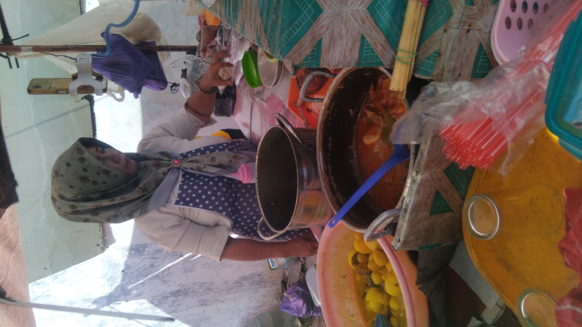 In Batang, Indonesia, a woman wearing a patterned hijab and a white top with a polka dot apron stands behind a makeshift food stall. She is in the process of serving or preparing food, with her right hand reaching towards a small white object. In front of her, on a table covered with a patterned cloth, are several cooking pots containing food. One pot holds a yellow curry-like dish with small, round pieces. Another pot contains a red stew with pieces of meat and vegetables. To the right of the pots, a stack of red plastic straws in a plastic bag is visible, along with a bamboo bundle tied with green string. Behind the woman, a purple plastic bag is hanging, and various other items are part of the stall's setup. The scene suggests a street food vendor operating outdoors.