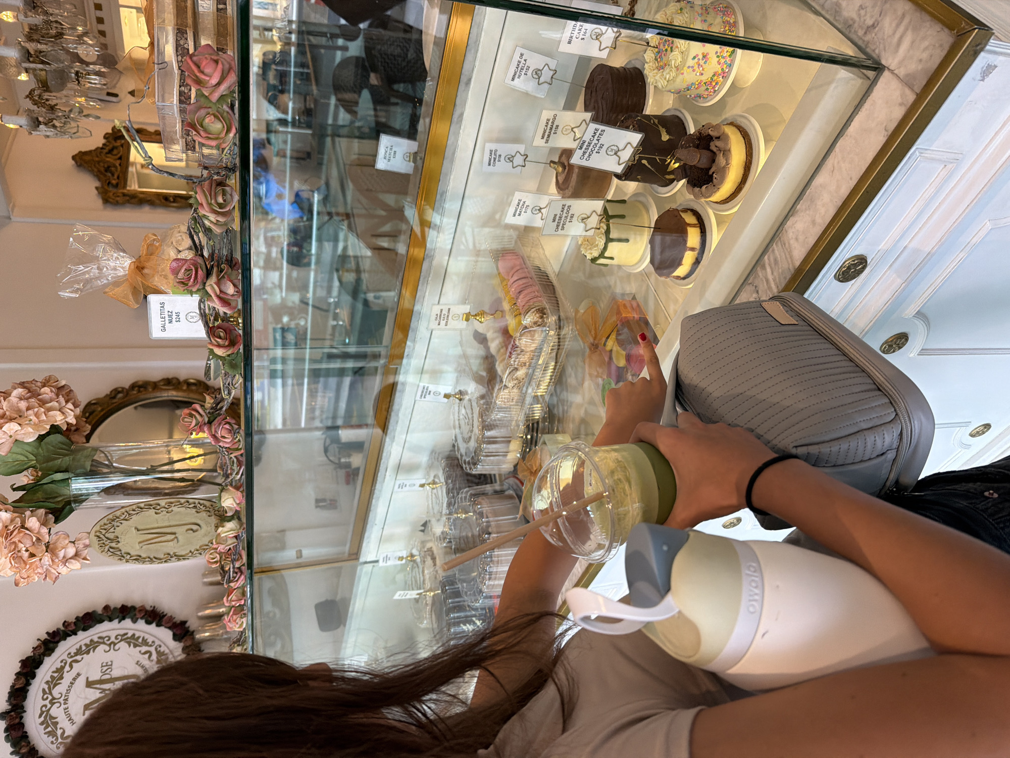 An indoor scene, likely a high-end patisserie or bakery in San Bernardino Tlaxcalancingo, Mexico, is captured from a slightly elevated perspective. In the foreground, the back of a person with long, dark hair is visible. They are holding a clear plastic cup with a green beverage and a straw in one hand, and a white and green Owala brand water bottle in the other. A gray quilted bag with a zipper is slung over their shoulder.
The person is positioned in front of a brightly lit glass display case filled with an array of meticulously presented desserts. The case showcases various mini-cakes, including Matcha, Conejito, Semiamargo, and Nutella, priced at $175, $158, $158, and $152 respectively. Also visible are Mini Cheesecakes (Speculoos $182, Chocolates $192), a Birthday Cake with sprinkles ($164), and a Rosca Matcha ($388). Individual "GALLETITAS NUEZ" (nut cookies) are also priced at $245.
The establishment's decor is elegant, featuring artificial pink roses draped along the edge of the display case, ornate gold-framed mirrors reflecting chandeliers, and an oval plaque bearing the letter "M". A circular sign partially visible in the lower left background reads "HAUTE PATISSERIE". The environment suggests a pleasant, perhaps indulgent, experience for patrons admiring or purchasing desserts. The lighting is bright, indicating daytime.