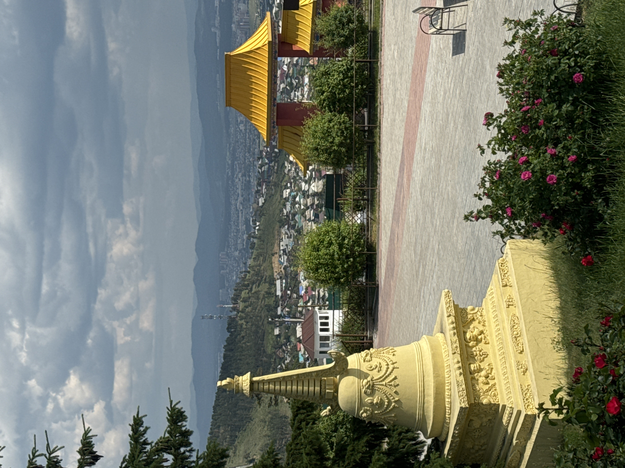 The image depicts a Buddhist stupa in the foreground, made of yellow material and adorned with intricate carvings. Behind the stupa, a paved walkway leads towards a complex of buildings with vibrant yellow roofs and red pillars, characteristic of Buddhist architecture. In the background, a sprawling cityscape of Ulan-Ude, Russia, unfolds, with a mix of residential buildings and distant hills. Green trees and bushes dot the landscape, and a cloudy sky looms overhead. A bench is visible in the middle ground, suggesting a public space. The overall scene appears to be a serene and picturesque vista overlooking the city.