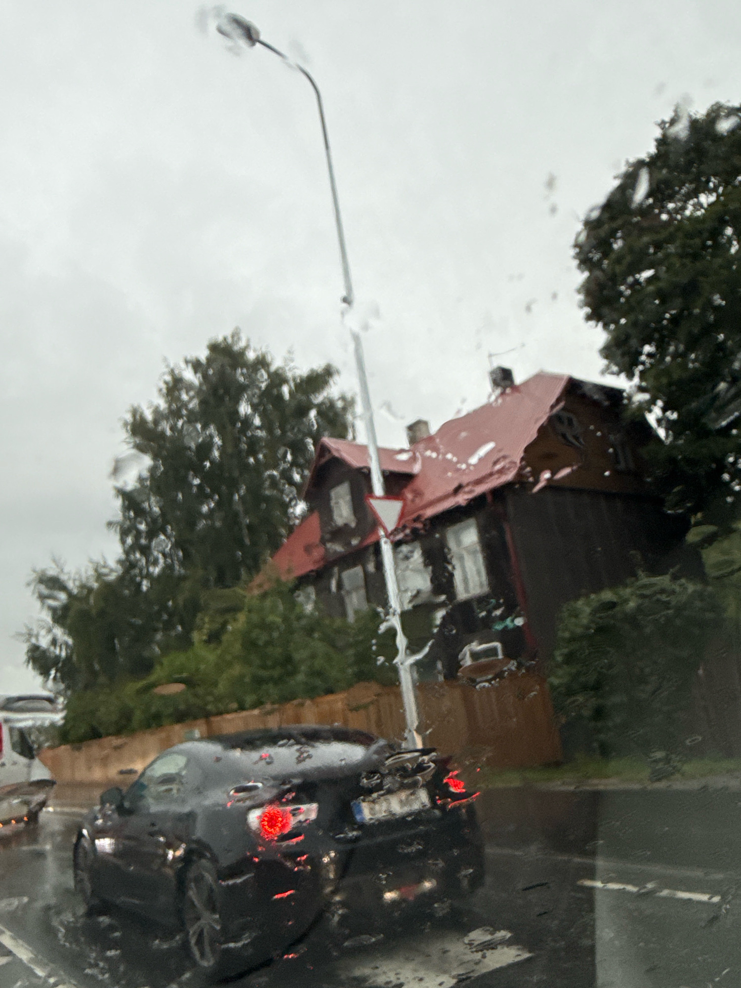 This is a medium-angle, slightly tilted, outdoor, daytime photograph taken through a wet window, likely from inside a vehicle, showing a street scene in Tallinn, Estonia. The scene is dominated by a dark-colored sports car with its brake lights illuminated, positioned in the foreground and slightly to the left. The car appears to be stationary or moving slowly in rainy conditions.
Behind the car, a traditional wooden house with a red roof and dark brown siding is visible. The house has several windows, some appearing to have curtains. A tall, grey street lamp stands to the left of the house, and a white triangular yield sign is attached to its pole. Trees with dark green foliage are scattered throughout the background, partially obscuring the house and the sky, which is overcast and grey.
The overall impression is of a gloomy, rainy day in a residential or mixed-use area of Tallinn. The presence of raindrops on the windowpane creates a slightly blurred and distorted effect, adding to the atmospheric quality of the image. The environment appears to be a street with wet pavement, indicating recent or ongoing rainfall. There are no visible people.