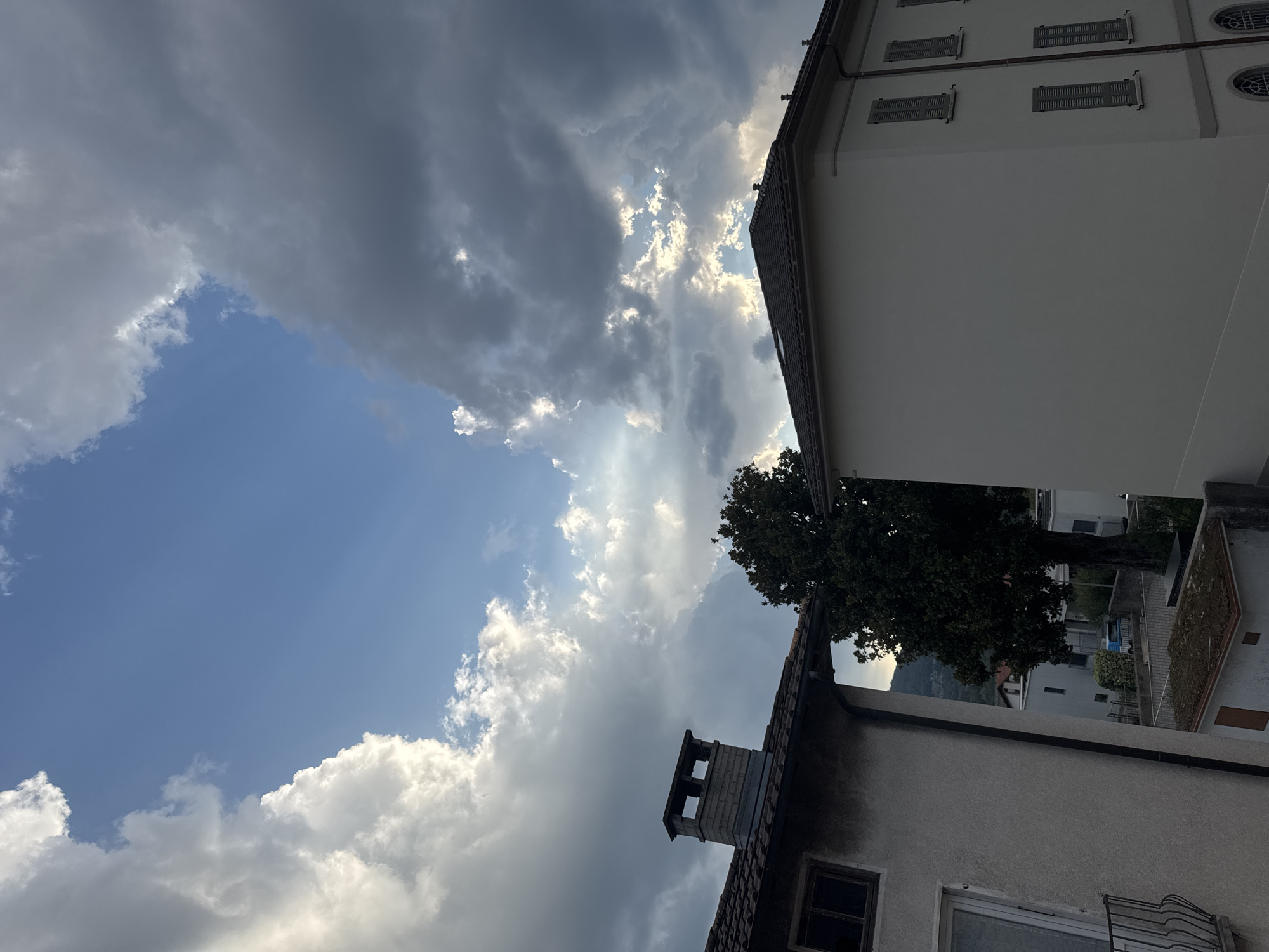 In Balerna, Switzerland, this image captures a view of buildings and a large tree against a dramatic sky. The sky is filled with a mix of light and dark clouds, with rays of sunlight breaking through in places. Two buildings are visible in the foreground. The building on the left is partially obscured and appears older, with a visible chimney and a tiled roof. The building on the right is a lighter color, with multiple windows that have closed shutters. A large, dark green tree stands between the two buildings. The background shows hints of rolling hills.
