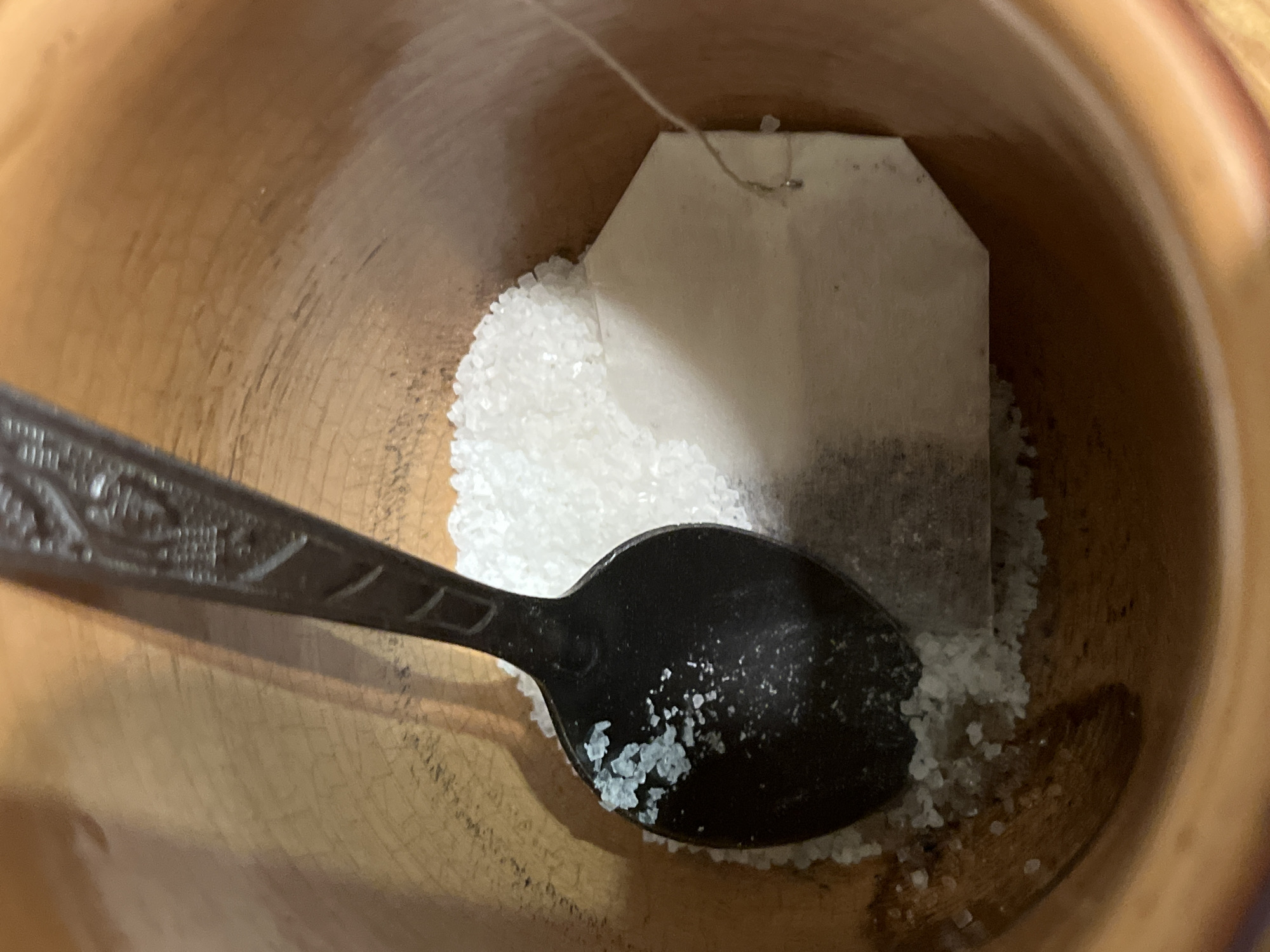 The scene is situated in Minsk, Belarus. The image captures a detailed, close-up, top-down perspective within a light brown, possibly ceramic or wooden, cylindrical container. The container's inner surface displays a subtle, irregular texture, suggesting either a material grain or a fine crackle finish. A dark-colored spoon, exhibiting decorative patterns on its handle, rests diagonally within the container. Its bowl is positioned over a significant accumulation of white, crystalline granules, indicative of sugar or salt. Small particles of this substance, some with a faint bluish tint, are observed adhering to the spoon's surface. Partially submerged in these granules and situated beneath the spoon's handle is a standard, rectangular tea bag. A thin, light-colored string extends upwards from the tea bag. The arrangement of objects suggests a static, preparatory state. No individuals are visible within the frame. The lighting indicates an overhead source, casting soft shadows.