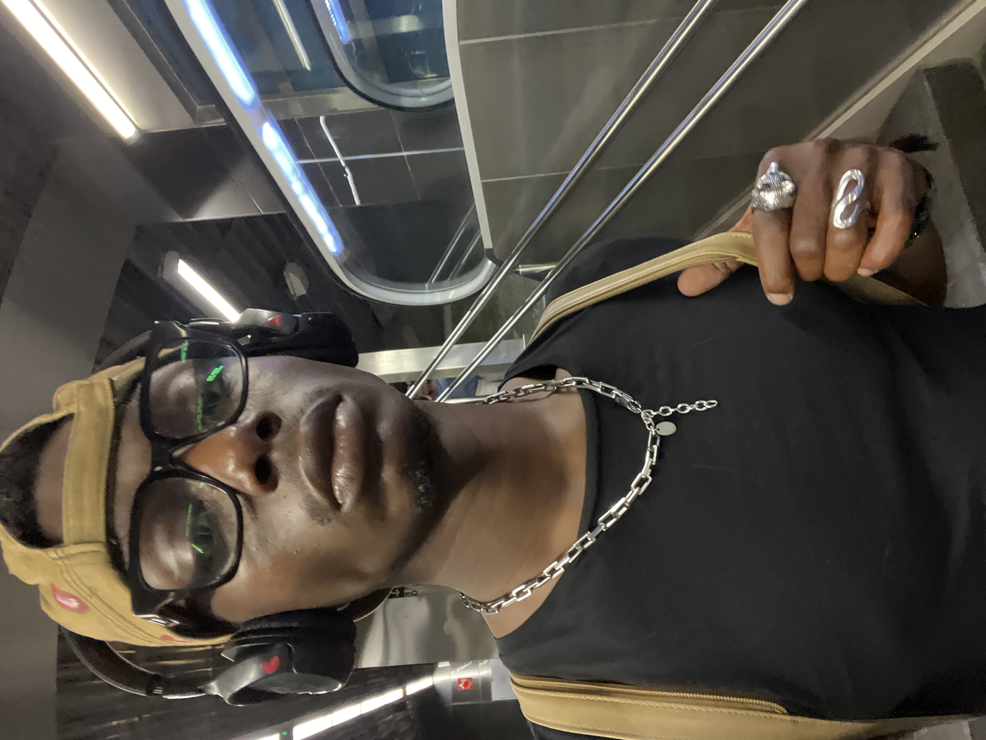 An individual with dark skin, wearing a backward tan baseball cap, black-rimmed glasses, and black over-ear headphones around his neck, is captured in a selfie-style shot. He has a neutral expression and looks directly at the camera. He is dressed in a black t-shirt and accessorized with a silver chain necklace that includes a small, circular pendant. His right hand, prominent on the right side of the frame, displays two distinctive silver rings: one appears sculptural and elaborate, while the other is an elongated, abstract loop design. A tan strap or zipper, possibly from a bag or backpack, is visible below his arm.
The setting is an indoor, modern public space, brightly illuminated by linear fluorescent light fixtures on the ceiling and walls. The background features sleek, metallic railings and reflective surfaces, suggesting glass or polished metal panels, consistent with contemporary architecture found in transportation hubs or public buildings. Given the context of Barcelona, Spain, this could be a metro station or a similar urban transit area. Reflections of green lights or screens are visible in the lenses of his glasses, and a faint red heart symbol can be seen on his headphones. The image is captured from a slightly upward angle, with the subject dominating the foreground.