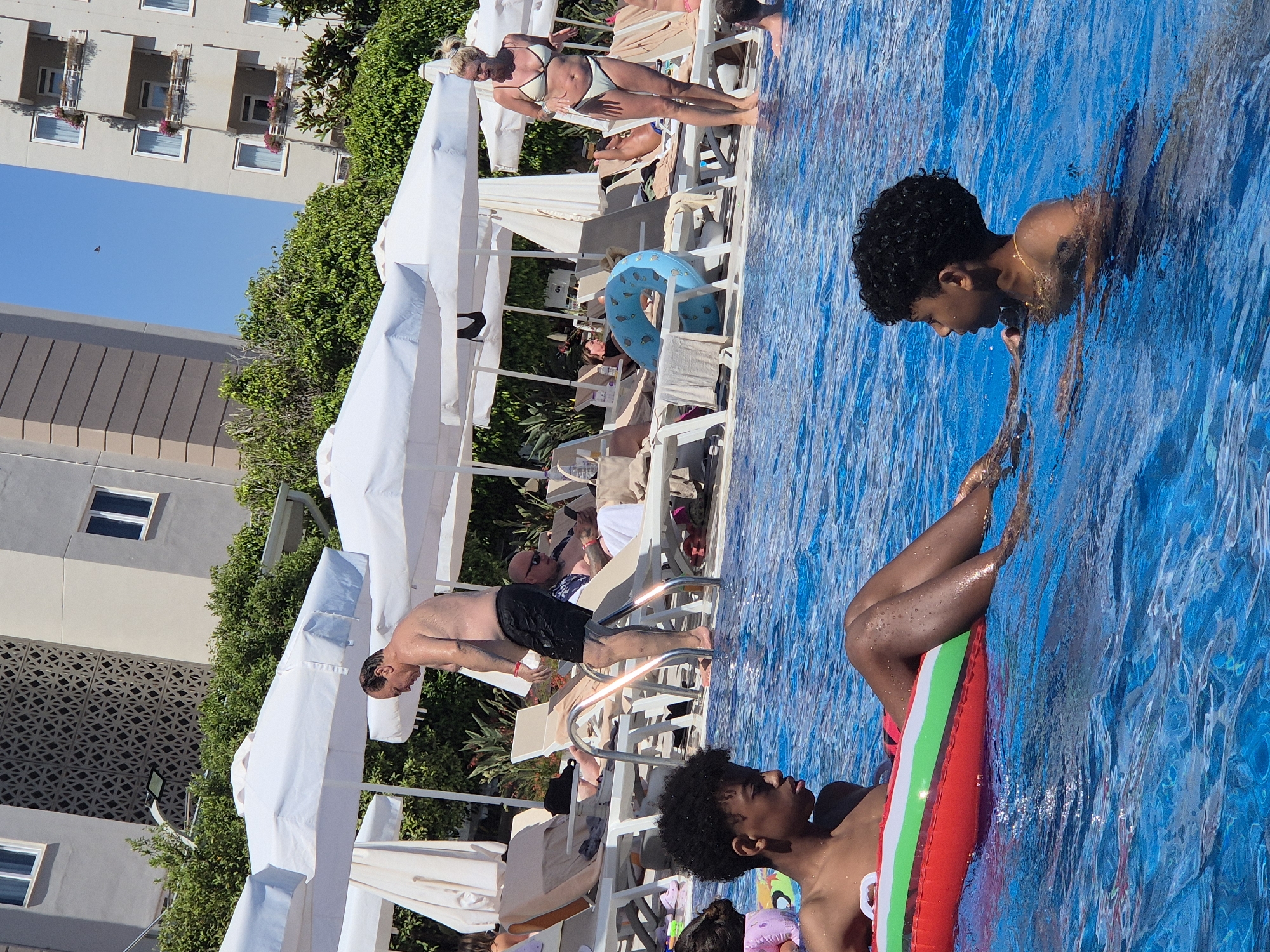 The image shows several people in and around a swimming pool on a sunny day. The water is a vibrant blue. In the foreground, two individuals are visible in the water. One is a person with dark, curly hair, looking downwards, with their body submerged up to their chest. They are wearing a necklace. The other is a person lying on their back on a colorful inflatable raft, with their legs bent. Their skin is dark, and they are partially submerged in the water.
In the background, several lounge chairs are arranged around the pool. On one of these chairs, a person in a white bikini is lying down, with their body partially in view. There are white umbrellas providing shade over some of the chairs. More people are seen relaxing on the lounge chairs. A blue inflatable ring with a pattern is visible on a chair.
Further in the background, a building with multiple windows and balconies is visible, surrounded by lush green trees. The scene appears to be a resort or hotel setting. The sun is shining brightly, indicated by the strong lighting and the clear blue sky. The overall mood seems relaxed and leisurely, typical of a vacation or a day at a swimming pool.