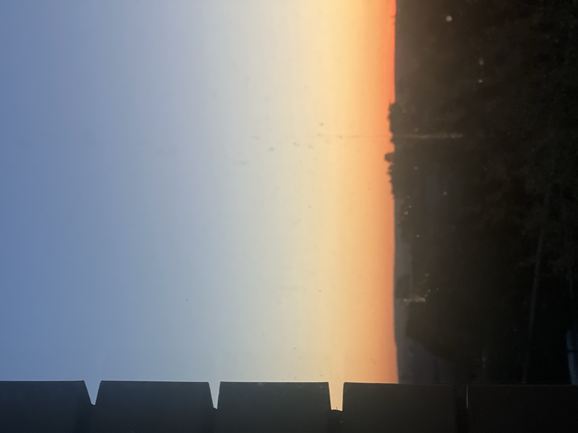 The image depicts a sunrise or sunset scene, possibly in Votkinsk, Russia. A gradient sky transitions from blue to orange hues. A dark, indeterminate object, likely vegetation or buildings, is on the right side. A dark, silhouetted structure with repeating rectangular cutouts is visible in the foreground.