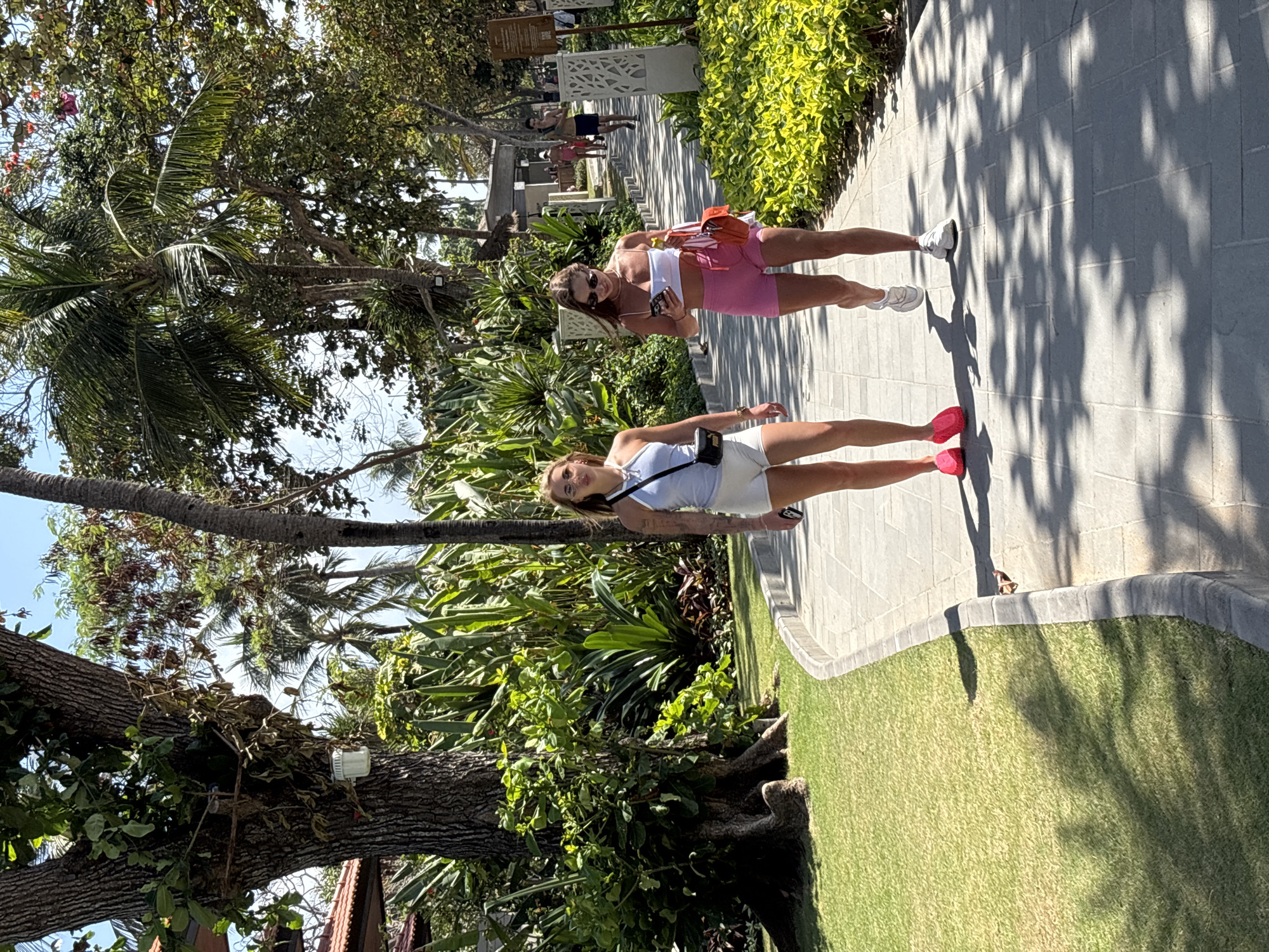 In Kuta Selatan, Indonesia, two young women walk along a paved pathway lined with lush green grass and dense tropical foliage. The woman on the left wears a light blue halter top and white shorts, with a black crossbody bag. The woman on the right wears a white bandeau top and pink athletic shorts, carrying a bright orange purse and holding a smartphone. Sunlight filters through palm trees, casting dappled shadows on the path. In the background, other individuals are visible, along with resort-like structures and more trees.