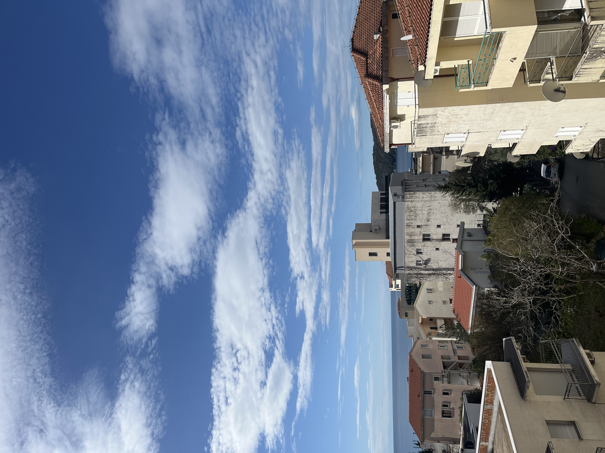 The image captures an outdoor daytime scene from a high vantage point in Boreti, Montenegro. Buildings with red-tiled roofs and light-colored exteriors occupy the right side of the frame. Balconies and satellite dishes are visible on some of the buildings. A street is seen winding through the buildings with a car parked on it. The background shows a sliver of the Adriatic Sea. The sky, taking up the left side of the image, is blue with streaky, cirrus-like clouds. No people are visible in the image.