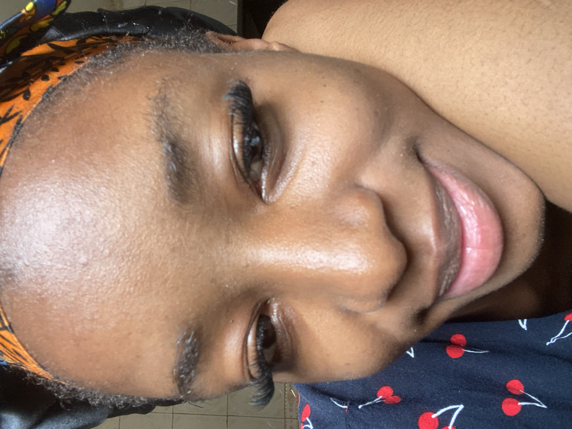 A close-up, eye-level portrait depicts an individual with a medium-dark skin tone, head tilted downwards and to the viewer's left. The person exhibits a closed-mouth smile, with the right side of their face appearing to rest on an unseen arm or hand.
The individual wears a black satin-like bonnet, revealing an orange, black, and yellow patterned fabric underneath at the hairline. Dark, tightly curled hair is visible at the temples. Prominent, long false eyelashes are applied to both upper eyelids, complementing dark brown irises. Eyebrows are dark and naturally shaped. Fine facial hair is present above the upper lip and on the jawline.
A dark-colored garment with a repeating pattern of red cherries and white stems is worn on the left side of the frame. The blurred background suggests an indoor environment with light-colored floor tiles visible in the upper left. No specific visual information in the image allows for confirmation of the location as Jos, Nigeria.