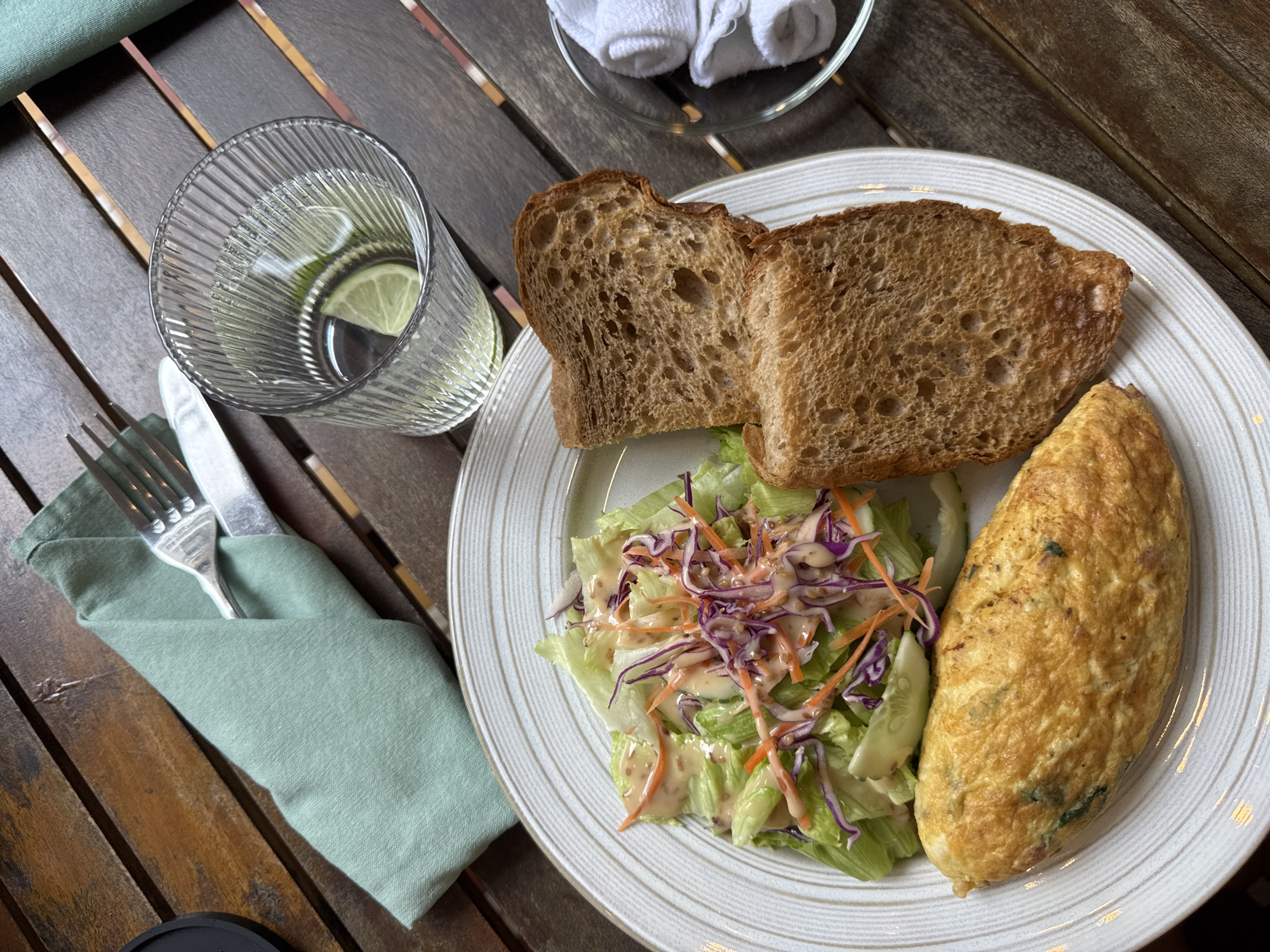A top-down shot of a plate of food and a glass of water on a wooden table, presumably in a restaurant or outdoor dining area. The plate contains two slices of whole-wheat toast, a salad with lettuce, shredded carrots, purple cabbage, sliced cucumber, and dressing, and an omelet. A green napkin holds a fork and knife. A glass of water with lime slices is to the left. A small dish with a white cloth napkin is in the upper right corner. The wooden table has alternating light and dark-stained planks. It is unknown if the location is in the city of Đà Nẵng, Vietnam.