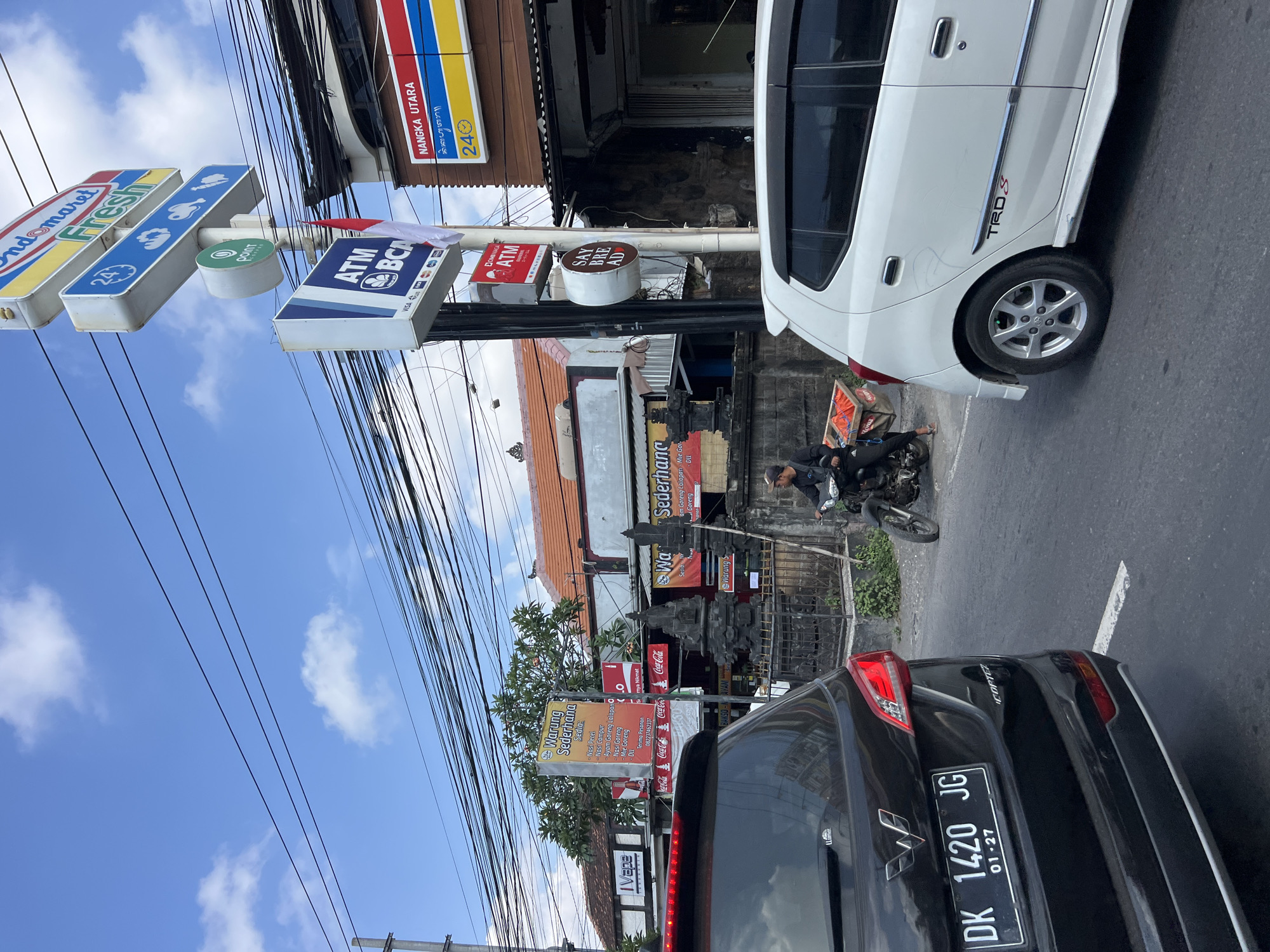 The image captures a street scene in Denpasar, Indonesia, on a bright, cloudy day. The foreground is dominated by the rear of a black SUV, displaying a license plate reading "DK 1420 JG". To the right, a white car is visible, with "TRD S" branding on its side.
A person on a motorcycle is in motion in the middle ground, with a wooden crate containing orange items attached to the back. Behind the motorcycle, a small, dark-colored shop front is partially obscured by a stone wall and decorative statues.
Several signs are prominently displayed. An "Indomaret Fresh" sign stands tall, with an ATM BCA sign below it. Another sign features the text "NANGKA UTARA" in blue and yellow lettering. A yellow sign for "Warung Sederhana" lists food items like "Nas Pree," "Ayam Goreng," and "Nas Goreng." Further to the left, a sign reads "IVape." Overhead, a complex network of electrical wires stretches across the sky. The overall environment suggests a bustling urban street with commercial establishments.
