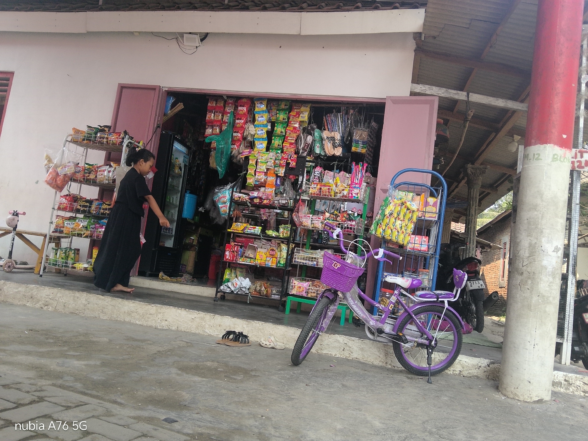 A medium shot shows a small shop with a woman standing inside, facing away from the camera and towards shelves stocked with goods. She is wearing a long black dress and barefoot. A purple bicycle with a basket is parked outside the shop on the right, angled towards the viewer. Several pairs of flip-flops are scattered on the ground in front of the shop and to the left of the bicycle. The shop appears to be a roadside stall, with an open front displaying merchandise. In the background, a motorcycle is parked. The scene is set on a concrete surface with some cobblestone detailing. The shop has a corrugated metal roof. The image is in color. This scene is located in Cakung, Indonesia.
