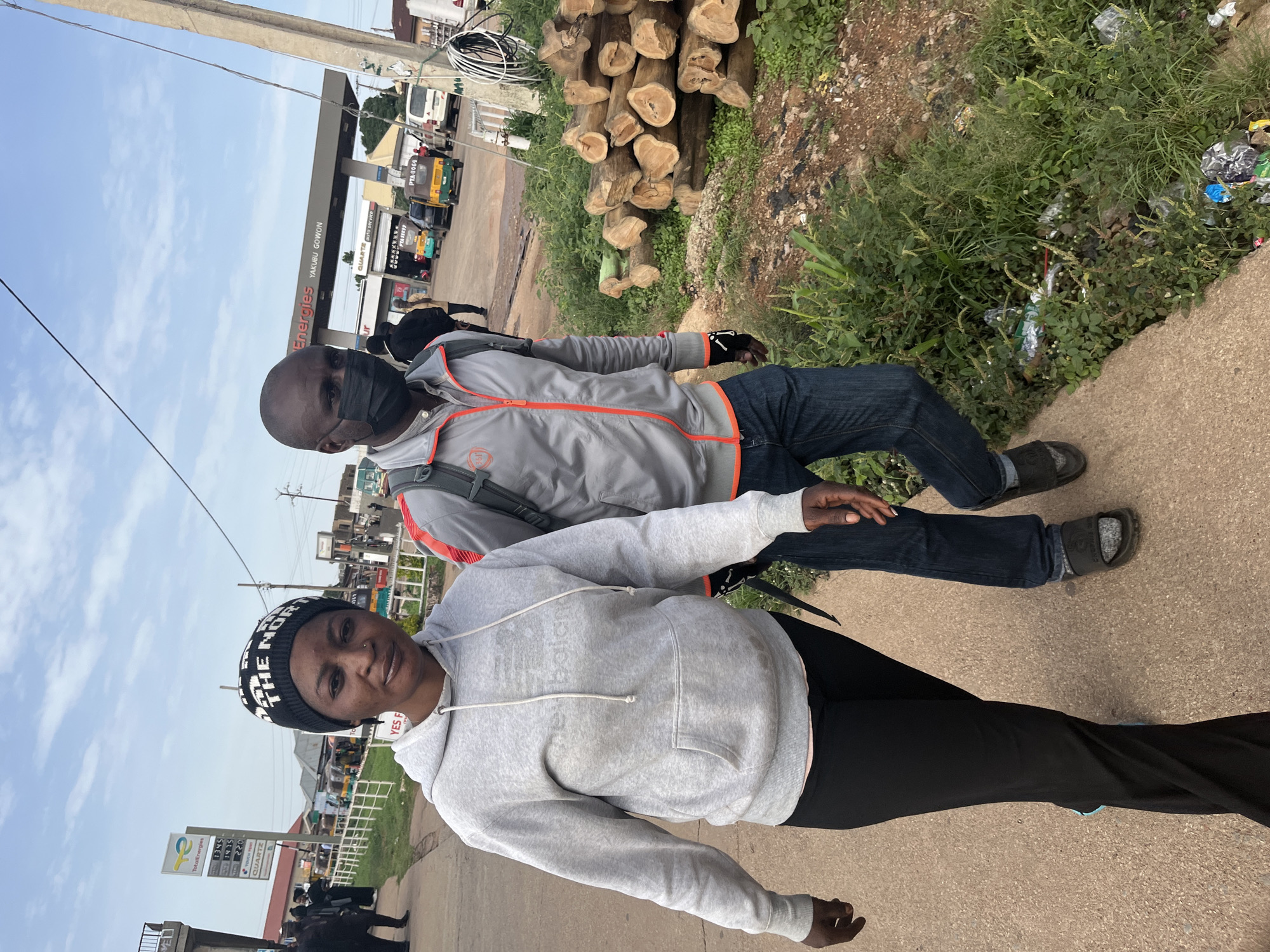 Two people are posing for a photo in front of a gas station in Gura Top, Nigeria. The person in the foreground, a woman, is wearing a grey hooded sweatshirt and a black and white beanie. She is smiling at the camera. The person behind her, a man, is wearing a grey jacket with orange accents, a black mask, and a backpack. They are both standing on a paved sidewalk.
In the background, the gas station has "Total Energies" and "Energies" visible on signs. There are also several vehicles, including what appear to be tricycles, at the gas station. To the right of the gas station, there is a pile of cut logs and an area with greenery and some litter. The sky is visible and appears to be a light blue, suggesting it might be daytime with clear weather. Text visible on signs includes "QUARTZ," "PTA-0064," and "YAKUBU GOWON." The overall scene suggests a casual moment captured at a roadside gas station.