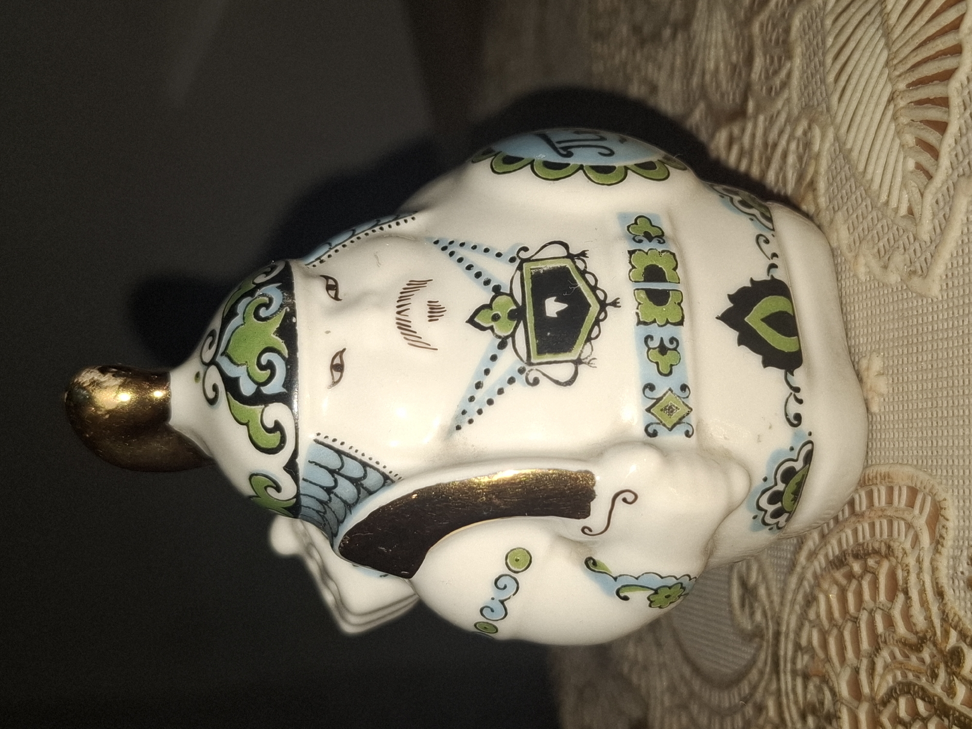 The image shows a close-up of a decorative ceramic figurine. The figurine is white with intricate hand-painted details in blue, green, and black, with accents of gold. It depicts a rotund figure wearing a helmet with a gold finial, adorned with scrolling green and blue patterns. The figure has a stylized face with thin eyes and a mustache. It wears a decorated tunic with a shield-like emblem on the chest and patterned bands around its waist and arms. One arm is raised, holding a golden object over its shoulder, possibly a staff or a bag. The figurine is placed on a textured, light-colored fabric surface, possibly a tablecloth, with a distinctive woven pattern. The background is dark and out of focus, suggesting controlled lighting for a product shot or still life. There is no indication of a specific location in Almaty, Kazakhstan, or any other city.