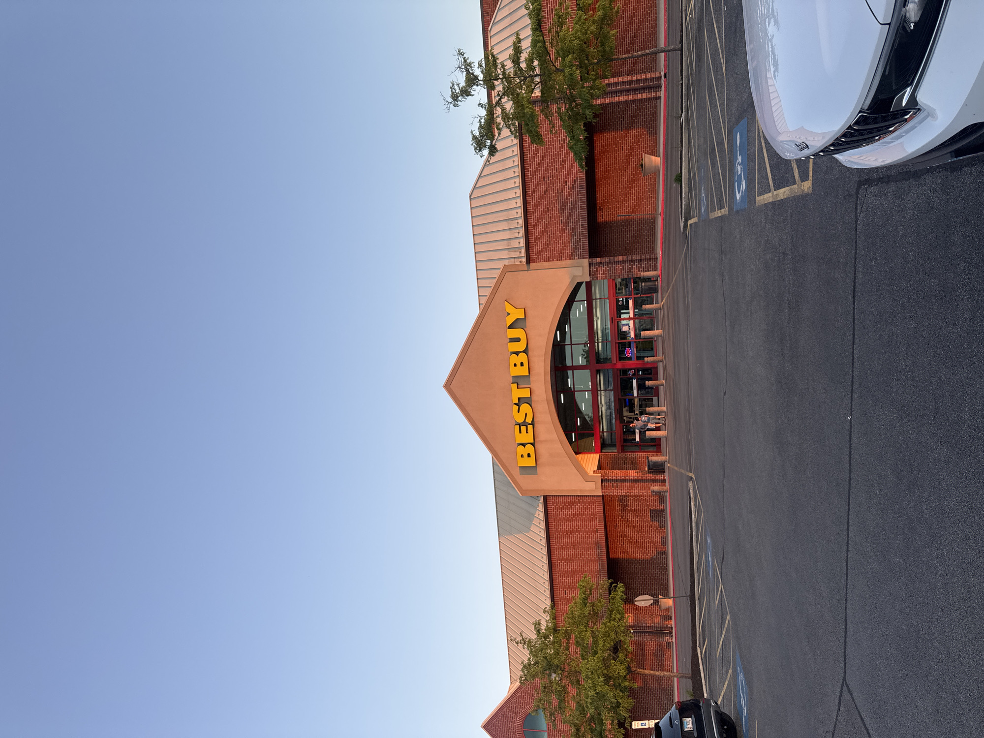 The image depicts the exterior of a Best Buy store in Bloomingdale, USA. The building has a brick facade with a distinctive triangular entrance. The yellow "BEST BUY" sign is prominently displayed. A parking lot is visible in front of the store, with a white car partially visible on the right edge. Trees are planted near the building. The sky is blue. There are people visible inside the store near the entrance. A handicap parking sign is visible in one of the parking spots.