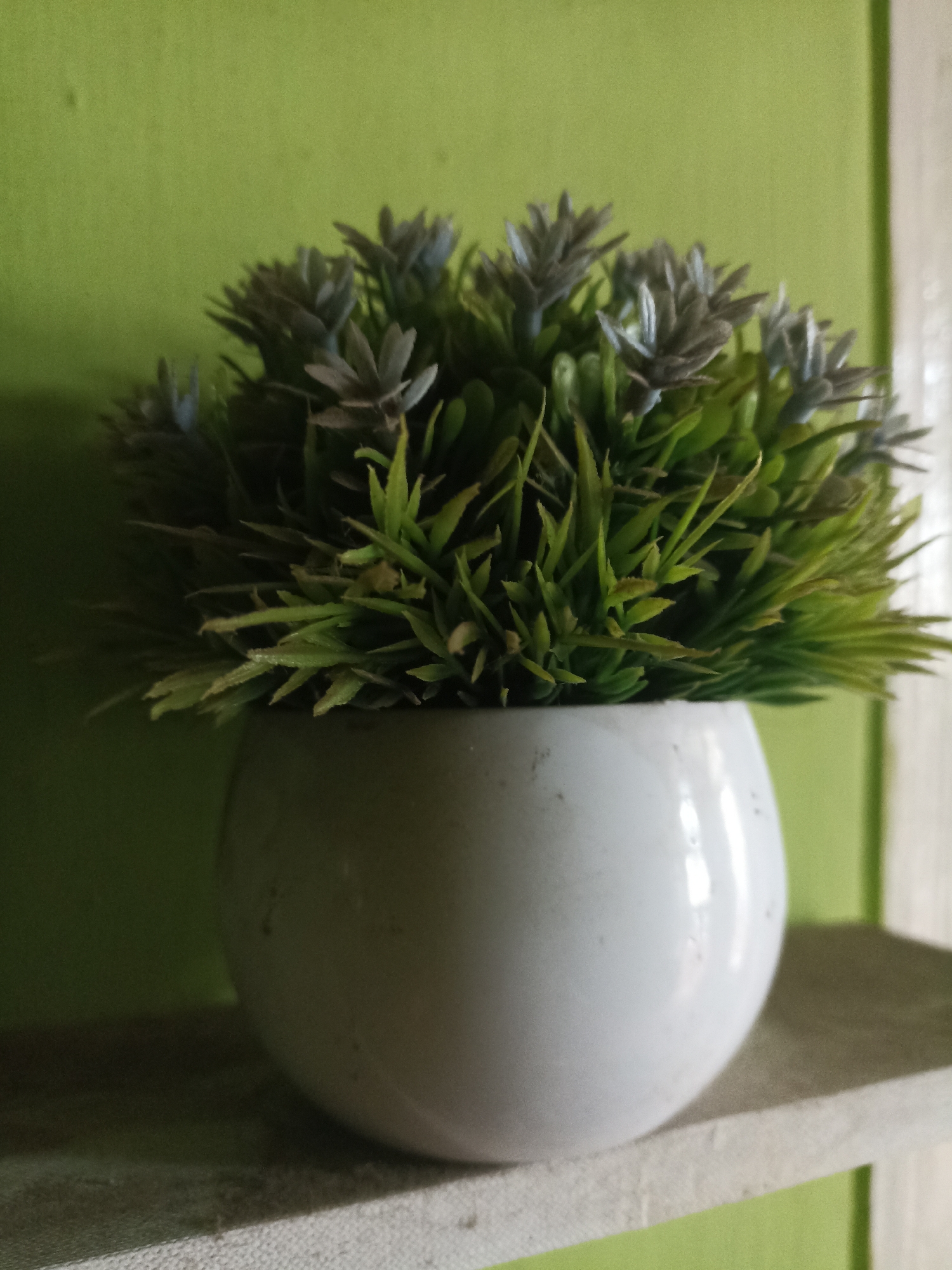 An artificial potted plant with green and grayish-blue foliage sits on a white shelf against a light green wall. The pot is white and spherical. The scene appears to be an indoor environment, possibly a home or office, located in Pandanarum, Indonesia. There are no people or animals visible. The lighting suggests it is daytime.
