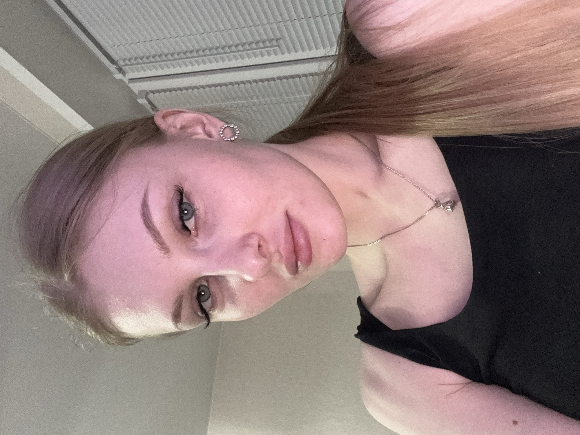 A light-skinned woman with long, light brown hair is seen from a low angle, facing the camera. She is wearing a black top and a delicate silver necklace. Her ears are adorned with small, circular earrings. The background includes a light gray wall and a white, slotted ceiling fixture. The location cannot be determined to be Perm, Russia, from the image alone.