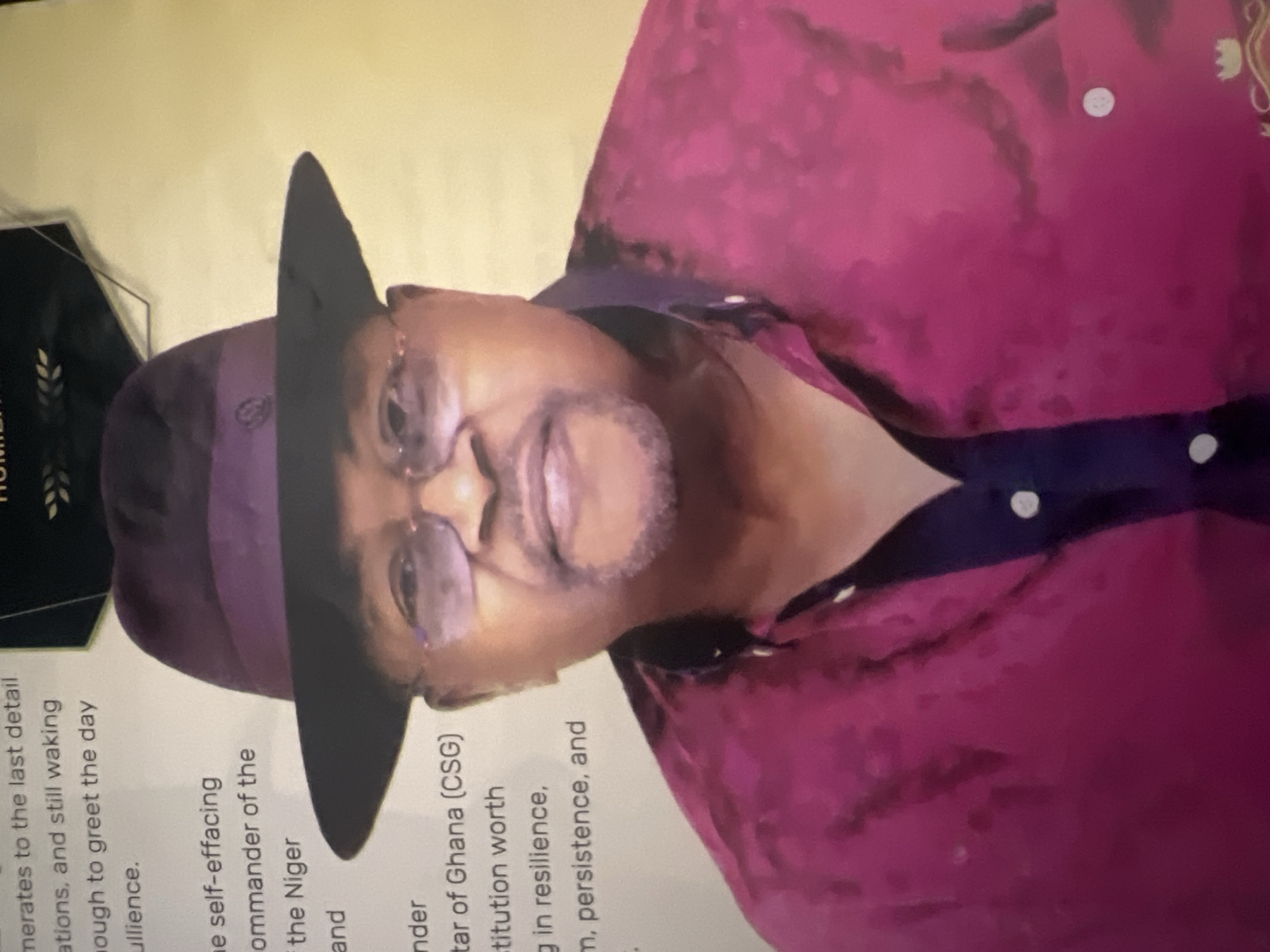 The image shows a headshot of a man wearing a purple fedora and a magenta collared shirt. He has on glasses and a goatee. He appears to be indoors, with a plain yellow background. Text is visible on the left side of the image, but it is out of focus and illegible. A dark framed award or certificate is visible at the top of the image. The image has no discernible connection to Jalingo, Nigeria, and does not depict any identifiable city or landscape.