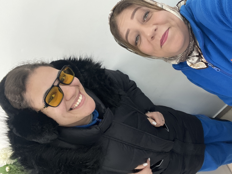Two women pose closely for a selfie. One wears a black winter coat with a furry hood and yellow-tinted sunglasses. She smiles broadly, revealing her teeth. The other woman wears a blue fleece jacket and a patterned headscarf. She smiles softly at the camera, her green eyes prominent. Both appear happy and comfortable. The background is a plain, light-colored wall.