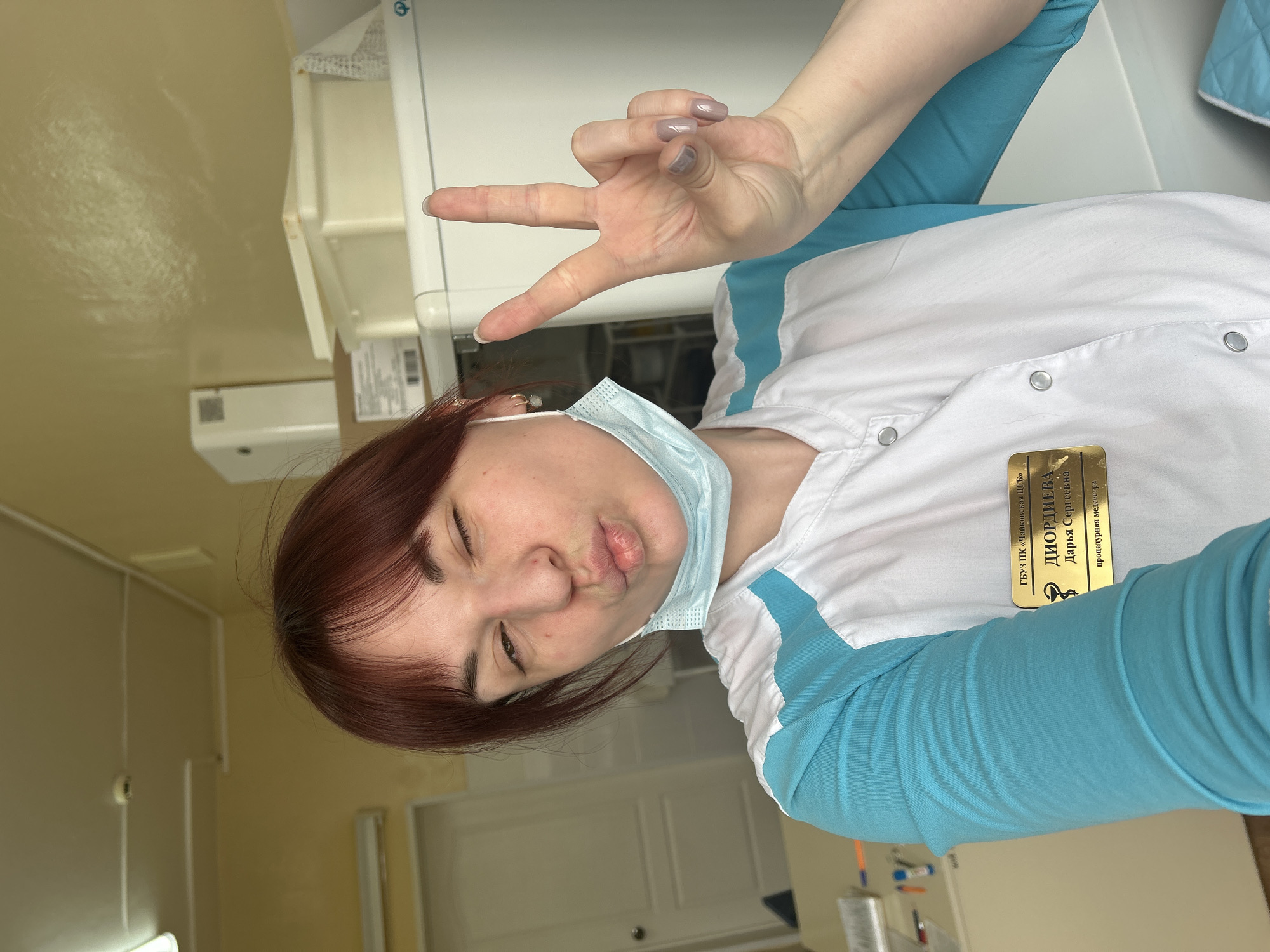 A woman in a white and blue uniform, wearing a surgical mask around her neck, is taking a selfie. Her hair is reddish-brown. She is making a peace sign with her left hand and puckering her lips. She is wearing a name tag written in Russian. The background includes a sterile environment with medical equipment and a pale yellow wall. The woman is located in the city of Фоки, Russia.
