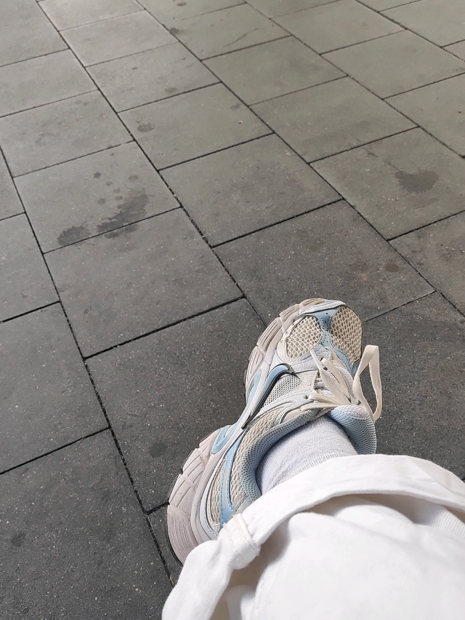The image shows a close-up, high-angle shot of a person's left foot, wearing a light blue and white athletic shoe with white laces and grey socks, partially obscured by white fabric. The foot is resting on a dark grey tiled pavement with visible grout lines and occasional dark spots, likely water stains. The scene is outdoors, on a paved surface, possibly a sidewalk or public square in the city of Ansbach, Germany. The lighting suggests it is daytime.