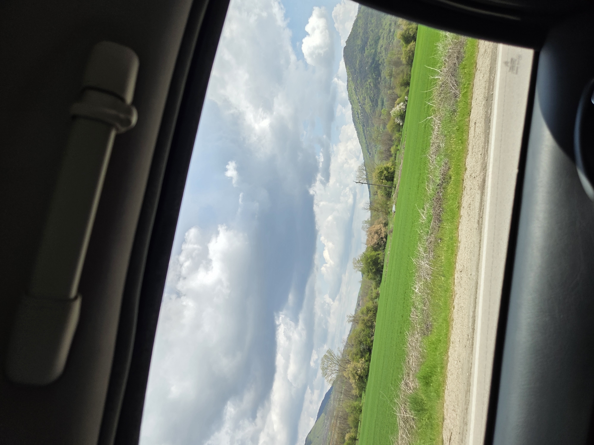 The view from inside a vehicle shows a landscape in Dobrodan, Bulgaria. A green field stretches towards a mountain covered in vegetation. A road runs along the edge of the field. The sky is cloudy with patches of blue. The interior of the vehicle includes a white grab handle.