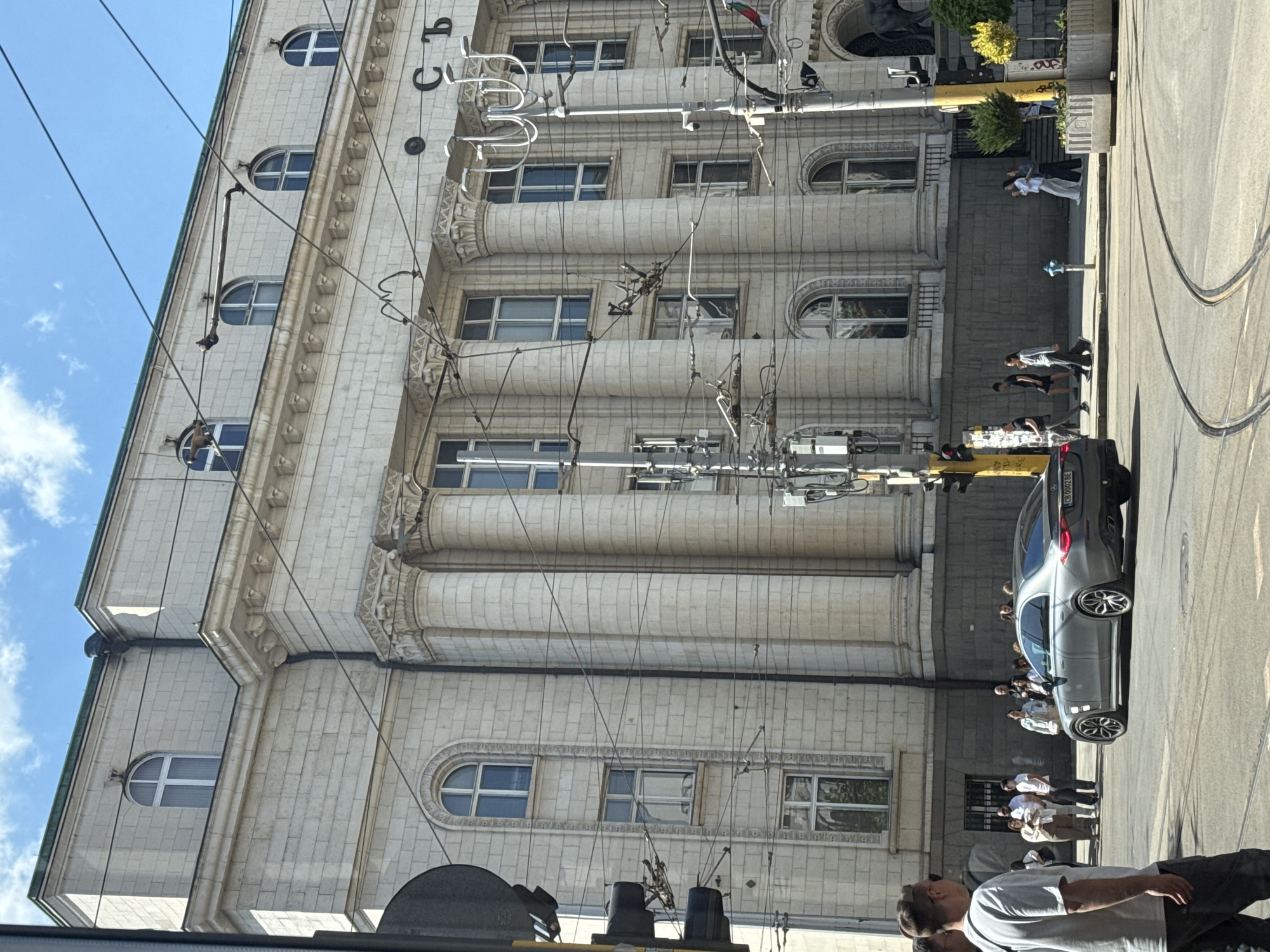 A street scene in Sofia, Bulgaria, displays a large, ornate building with rows of arched windows, imposing pillars, and detailed architectural moldings. Overhead tram lines and electrical cables crisscross the view. Pedestrians populate the sidewalk, some standing and observing. A silver car is parked near the building's facade. The sky is visible, suggesting a sunny day.