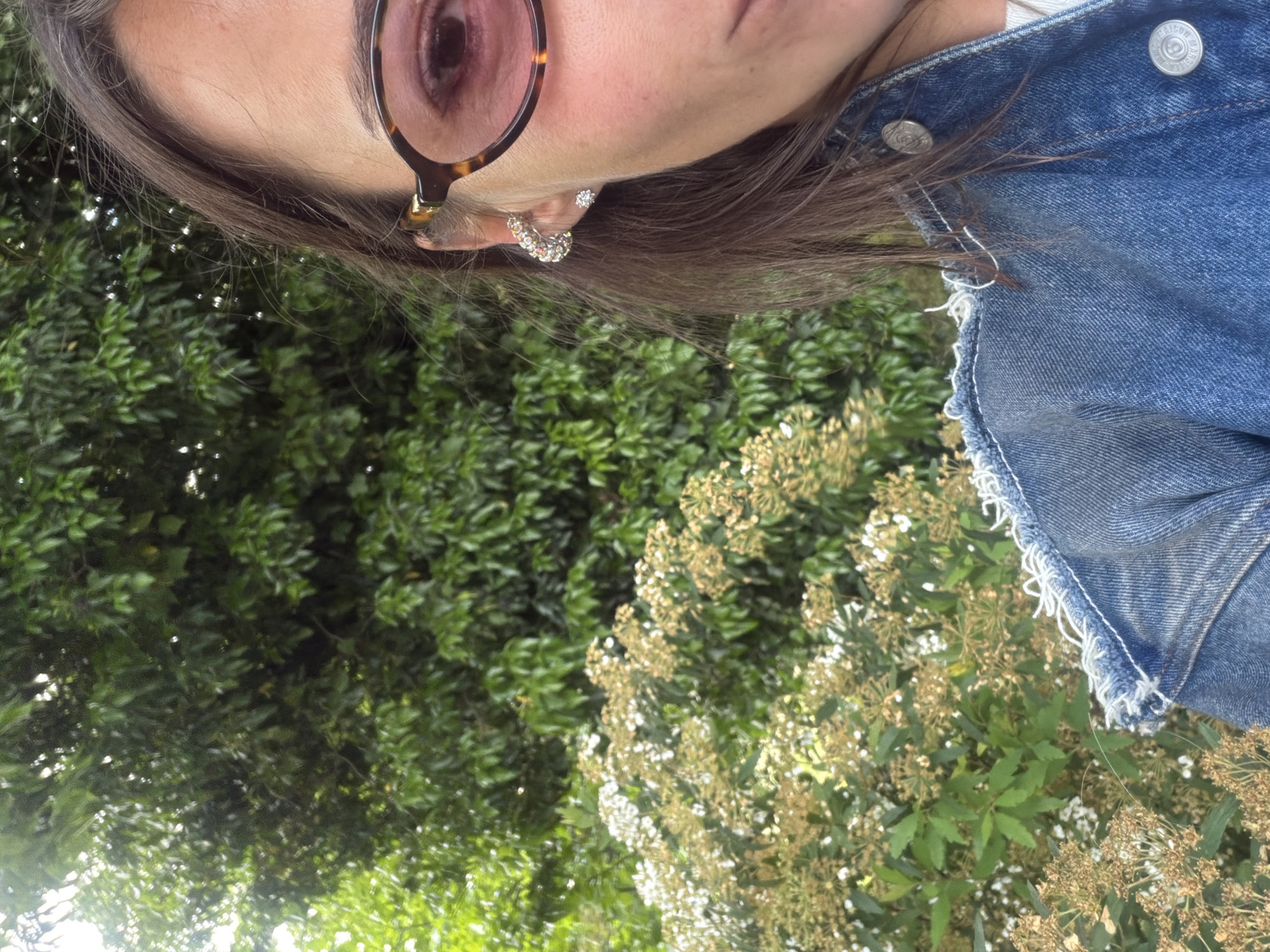 A close-up, angled shot captures a person wearing tortoise shell glasses and a denim jacket. The subject is positioned in front of lush green vegetation and clusters of small, white and tan flowers. Two earrings are visible in the subject's ear, one appears to be a small stud, and the other a larger, curved design. The environment suggests an outdoor setting. The location is stated as Napareuli, Georgia.